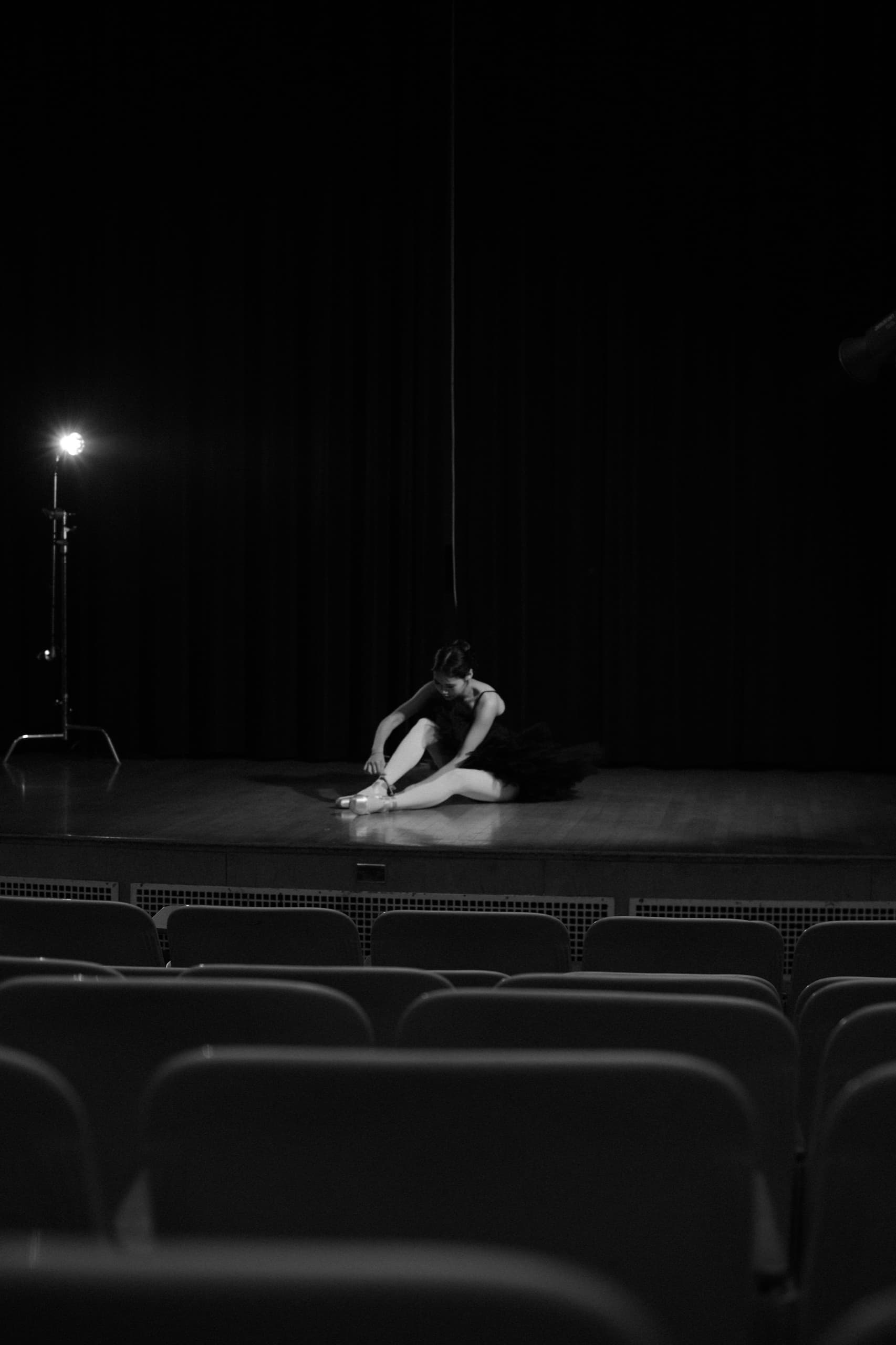 Black and white: a performer alone at the edge of a dark stage under a single hanging light, empty seats in the foreground.