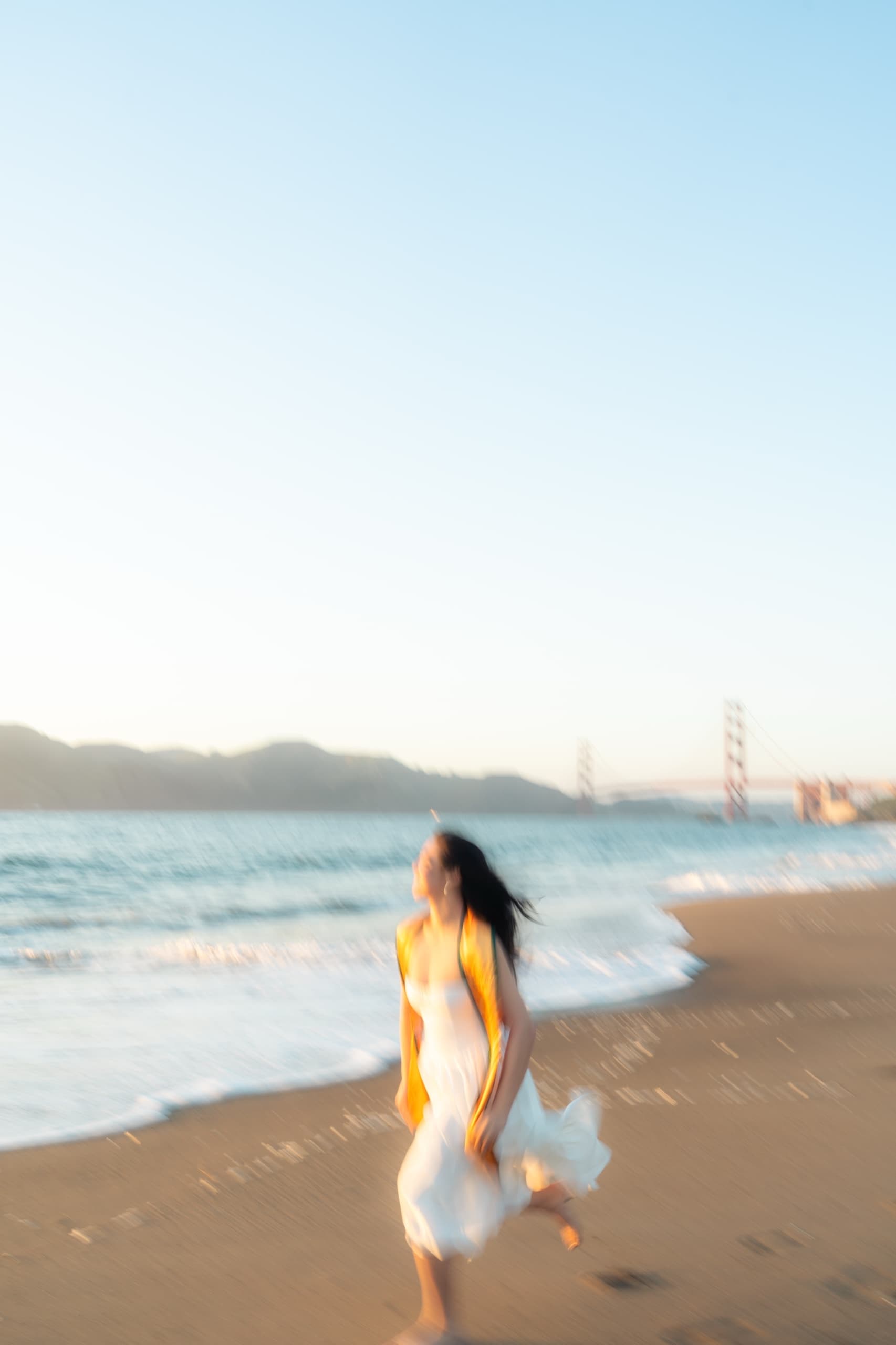 A graduate in a white dress and gold stole moves barefoot along the surf at sunset, the entire frame smeared by a long exposure, with the Golden Gate Bridge faint in the distance.