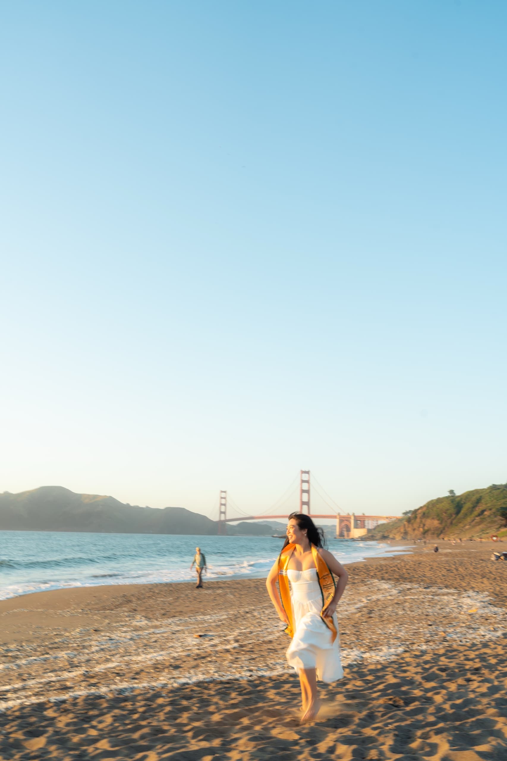A graduate in a white dress and gold honor stole runs barefoot across wet sand at sunset, with the Golden Gate Bridge and the headlands behind her under a wide, clear sky.