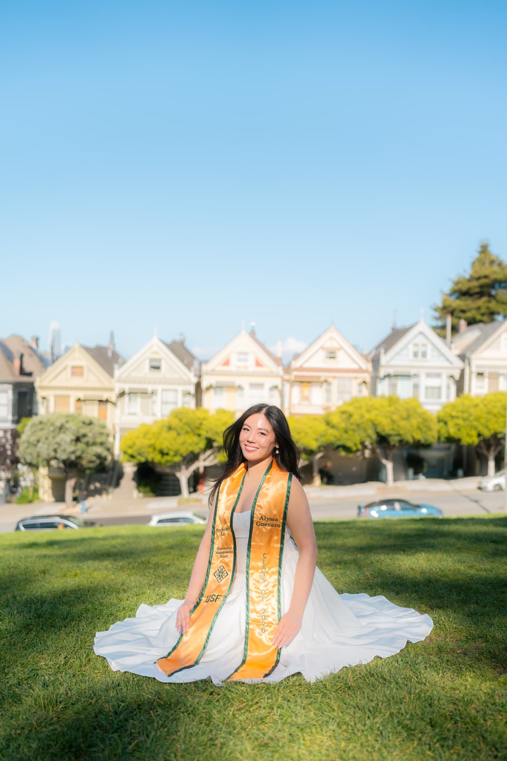 A graduate kneels on a sunlit lawn with the skirt of her white dress spread in a wide circle around her, wearing a gold University of San Francisco Class of 2026 stole, with the row of Victorian houses at Alamo Square behind her under a clear blue sky.
