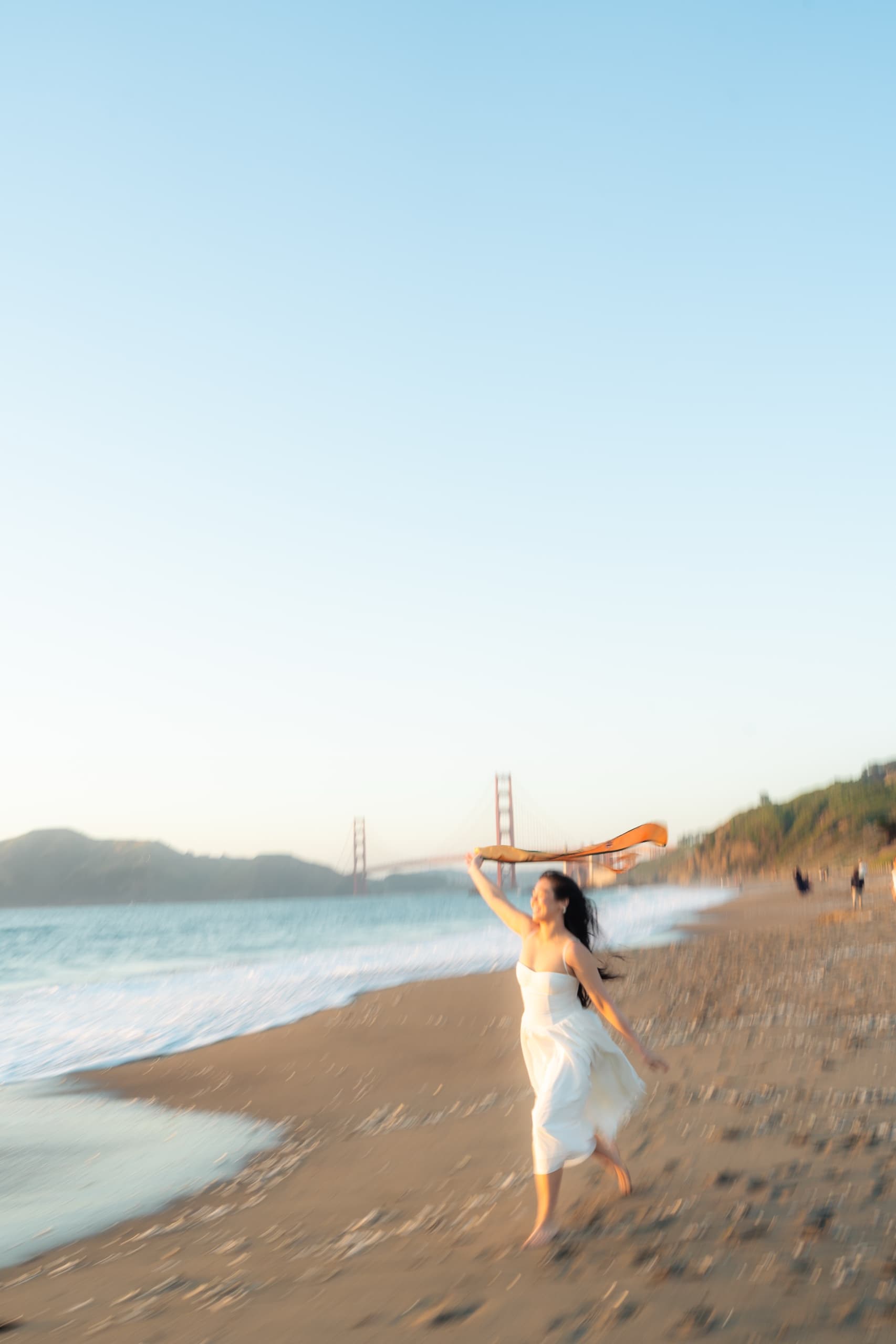 A graduate in a white dress runs barefoot along the beach holding her gold honor stole stretched above her head, motion blur streaking the sand and surf, with the Golden Gate Bridge behind her.