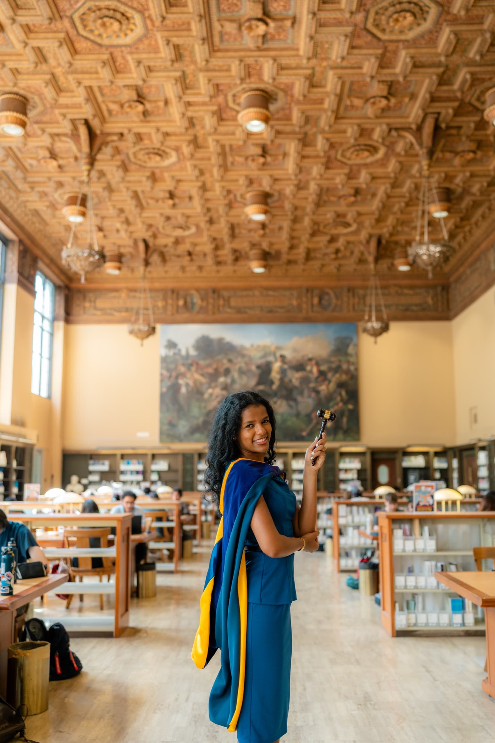 A law graduate in a blue suit turns to smile over her shoulder while raising a wooden gavel, a blue and gold academic hood over her arm, standing in the center aisle of a grand library reading room with a carved wood ceiling, hanging lamps, a large painted mural, and students working at long tables behind her.