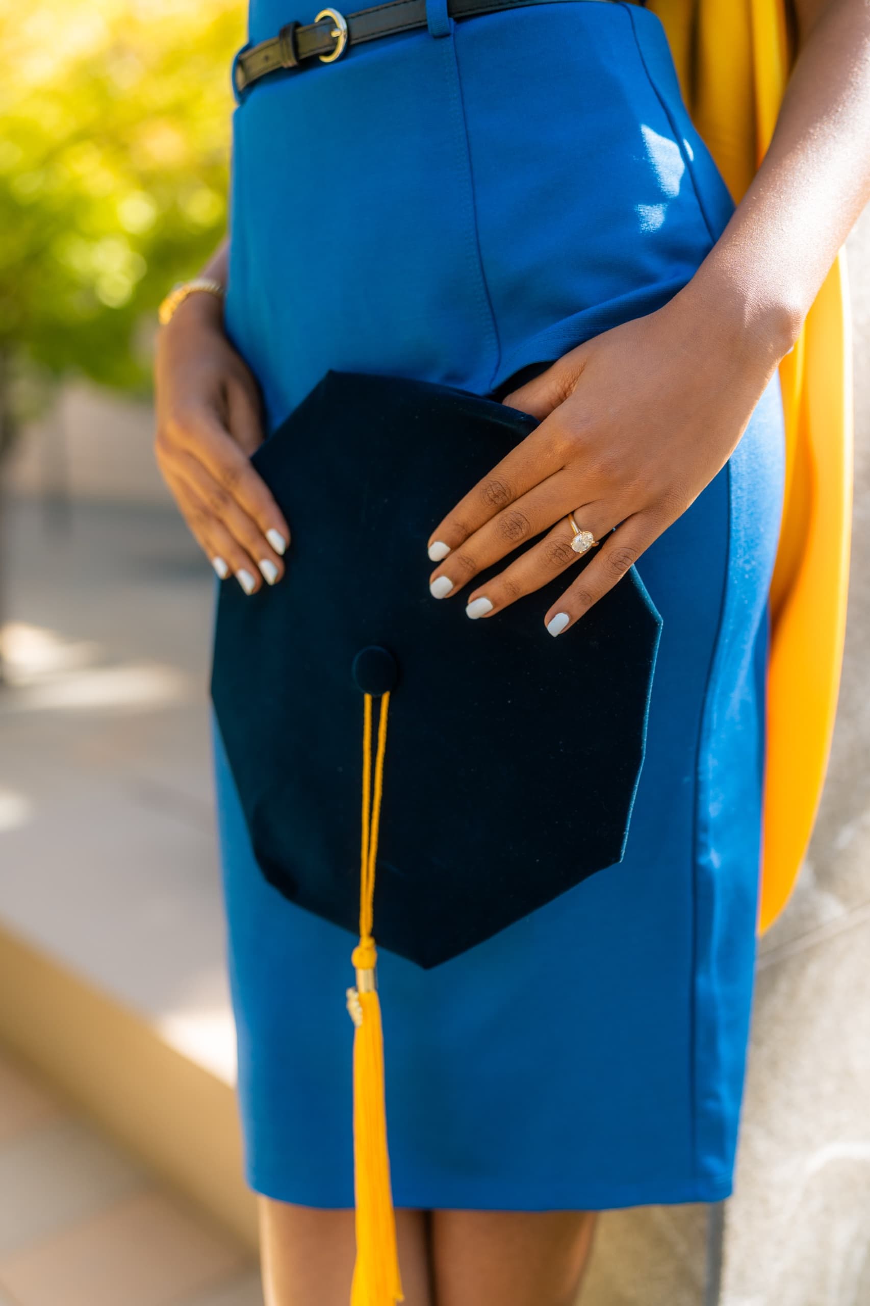 Close-up of a graduate's hands resting on a navy velvet graduation tam with a gold tassel, held against her cobalt blue dress, an engagement ring on her left hand and a gold academic hood draped over her shoulder in warm afternoon light.