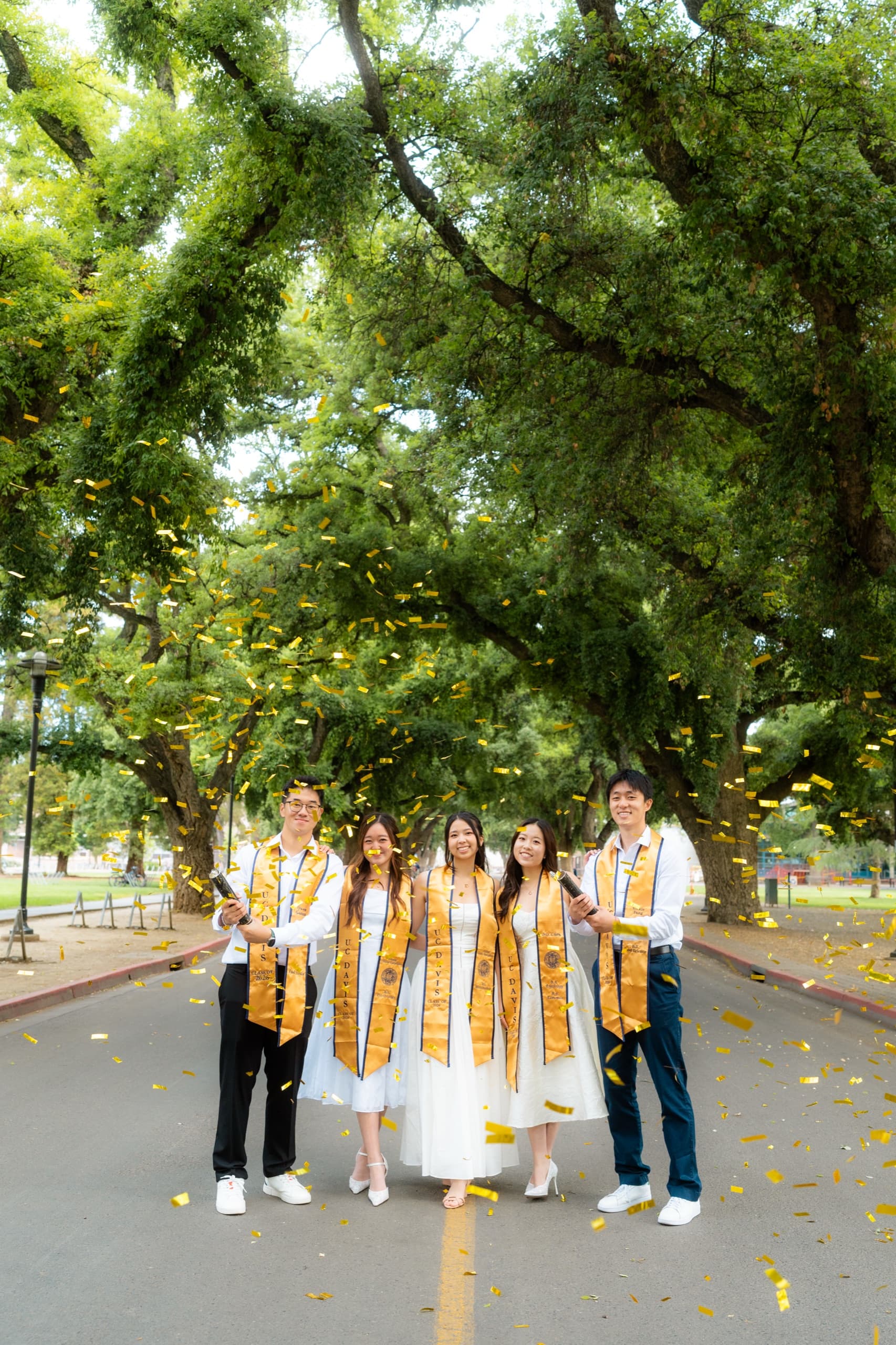 Five UC Davis graduates in white dresses and white shirts with gold stoles stand shoulder to shoulder on a tree-lined campus road while gold confetti falls around them from poppers held at each end of the line.
