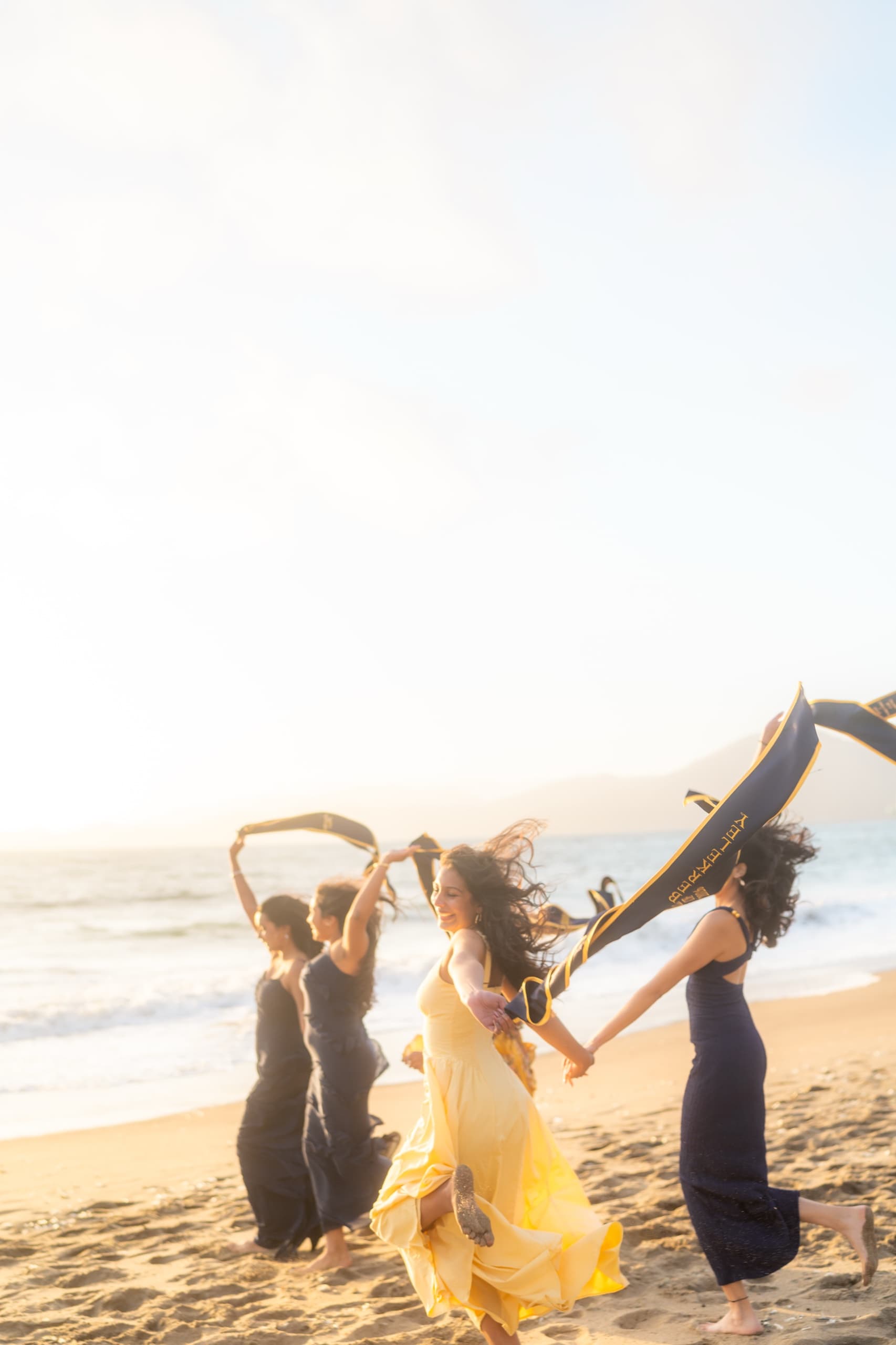 Four graduates in long dresses run barefoot along wet sand at sunset, holding navy and gold graduation stoles above their heads so the fabric streams out behind them, with breaking waves and hazy coastal hills in the background.