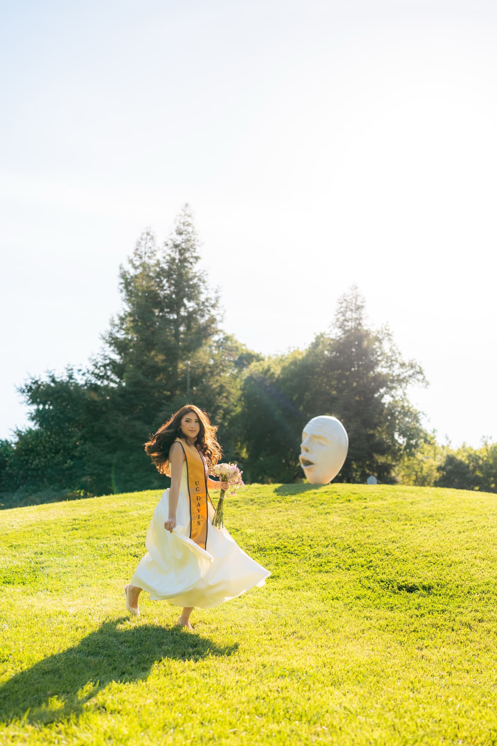A UC Davis graduate in a white dress and gold stole turns on a sunlit grassy hill holding a bouquet of pink and white flowers, her skirt swinging out, with a large white sculpture of a face on the lawn behind her and backlit trees along the ridge.