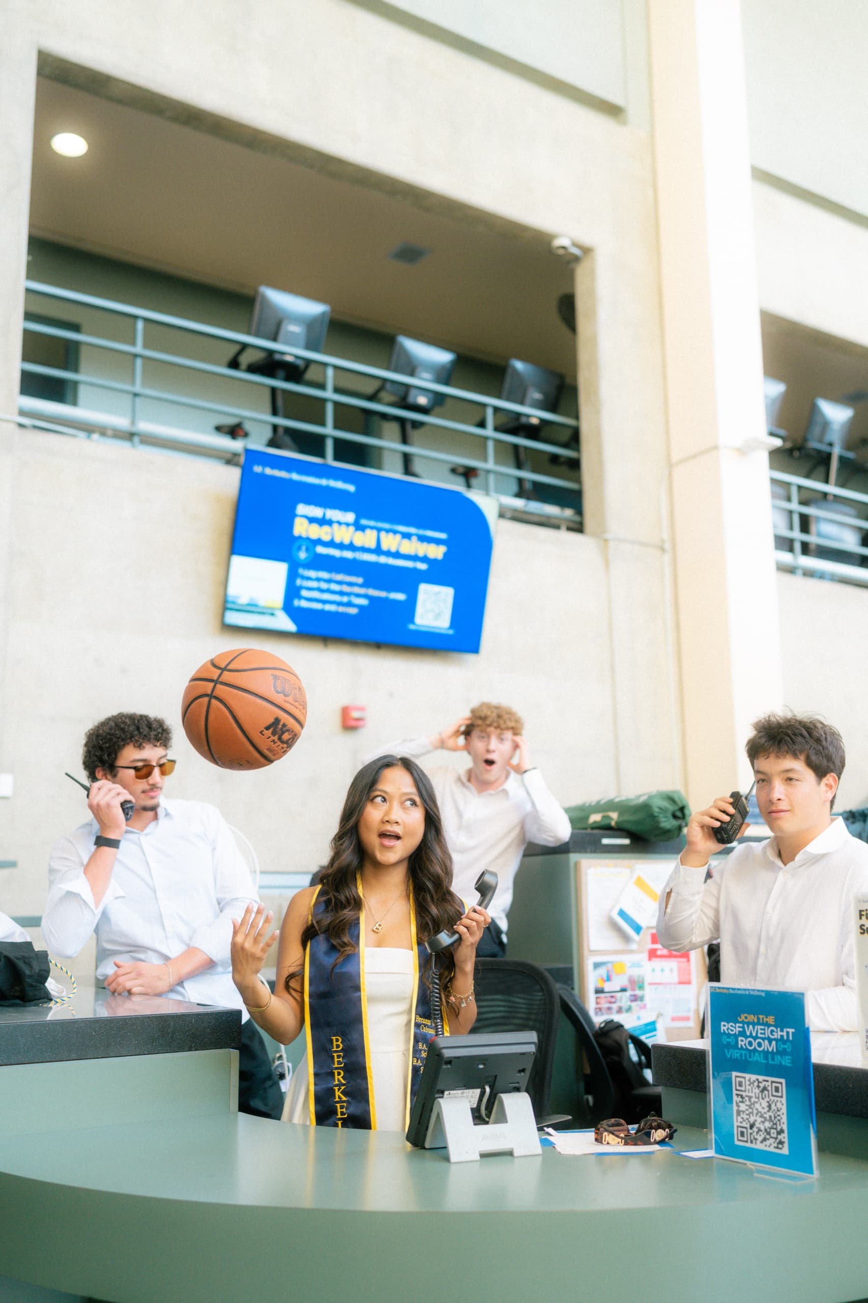 A graduate in a white dress and navy Berkeley stole stands behind a campus recreation center front desk, tossing a basketball into the air while holding a phone receiver, as three coworkers in white shirts react around her.