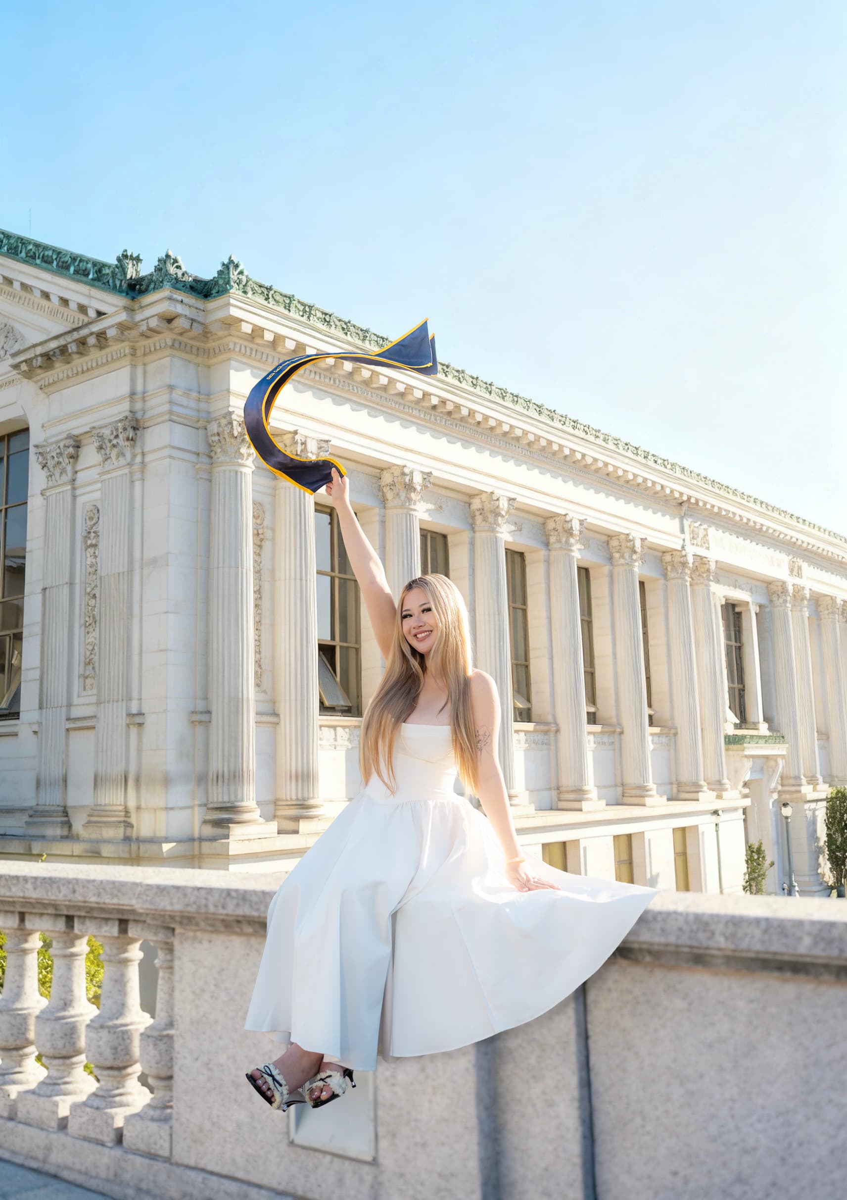 A graduate in a white strapless dress sits on a stone balustrade in bright sunlight, holding a navy and gold graduation cap high overhead with the tassel flying, in front of a columned neoclassical campus building.