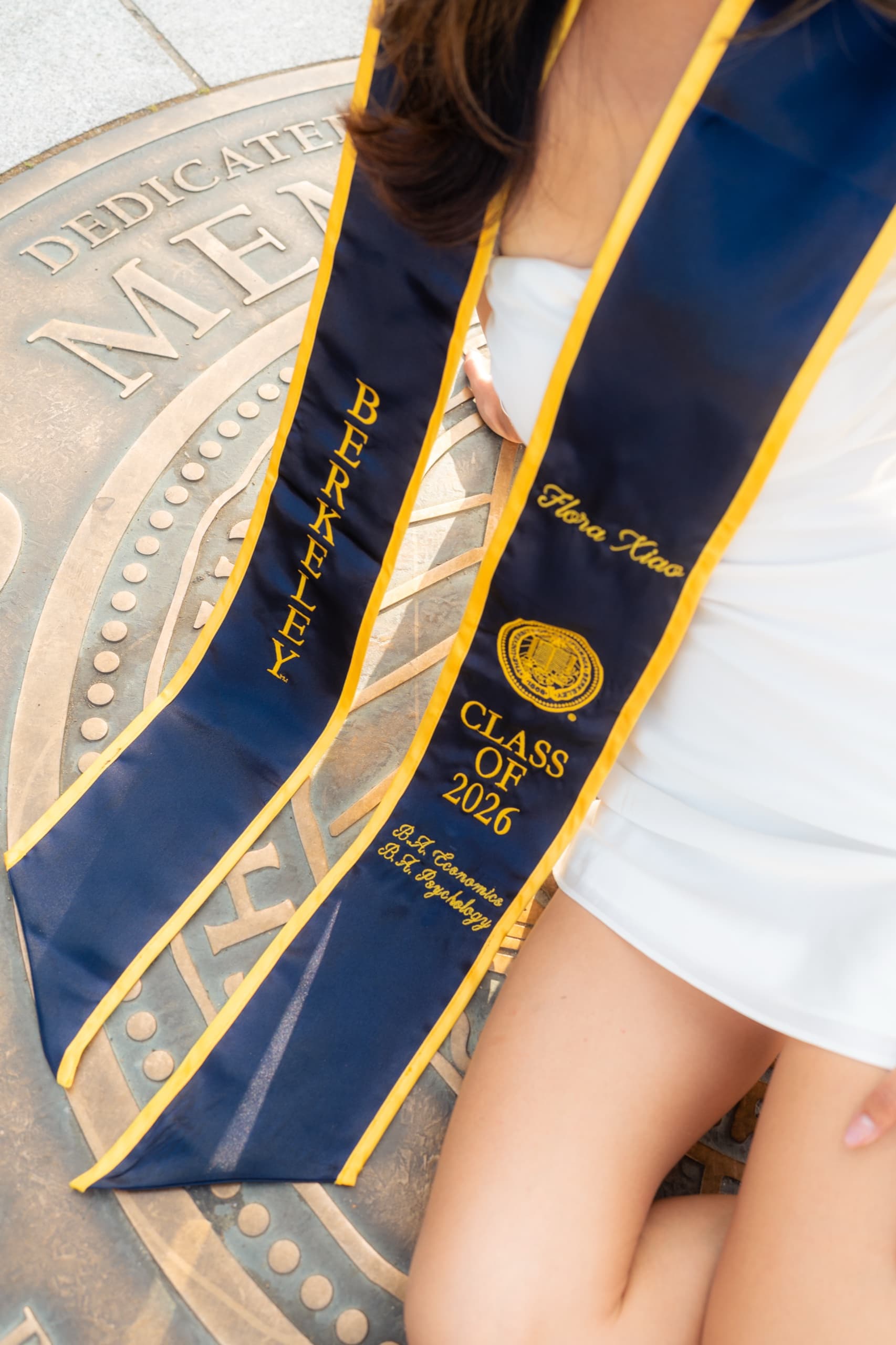 Overhead detail of a navy and gold graduation stole embroidered with the name Flora Xiao, Class of 2026, and two majors in economics and psychology, draped across a bronze university seal set into the pavement, with the graduate's white dress and hand at the edge of the frame.