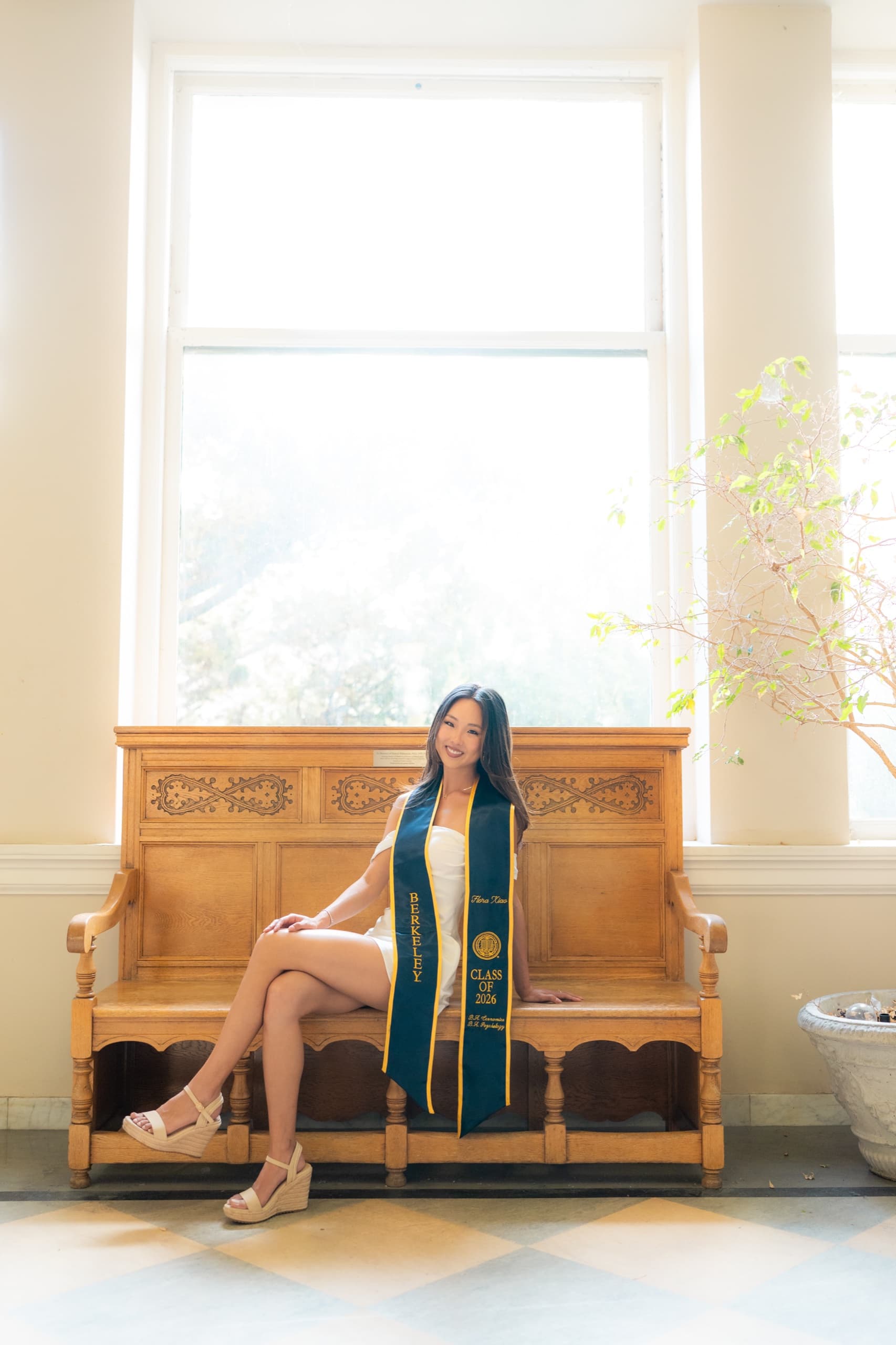 A graduate in a white dress and navy Berkeley stole sits with her legs crossed on a carved oak bench in front of a tall bright window, on a checkered tile floor with a potted tree to her right.