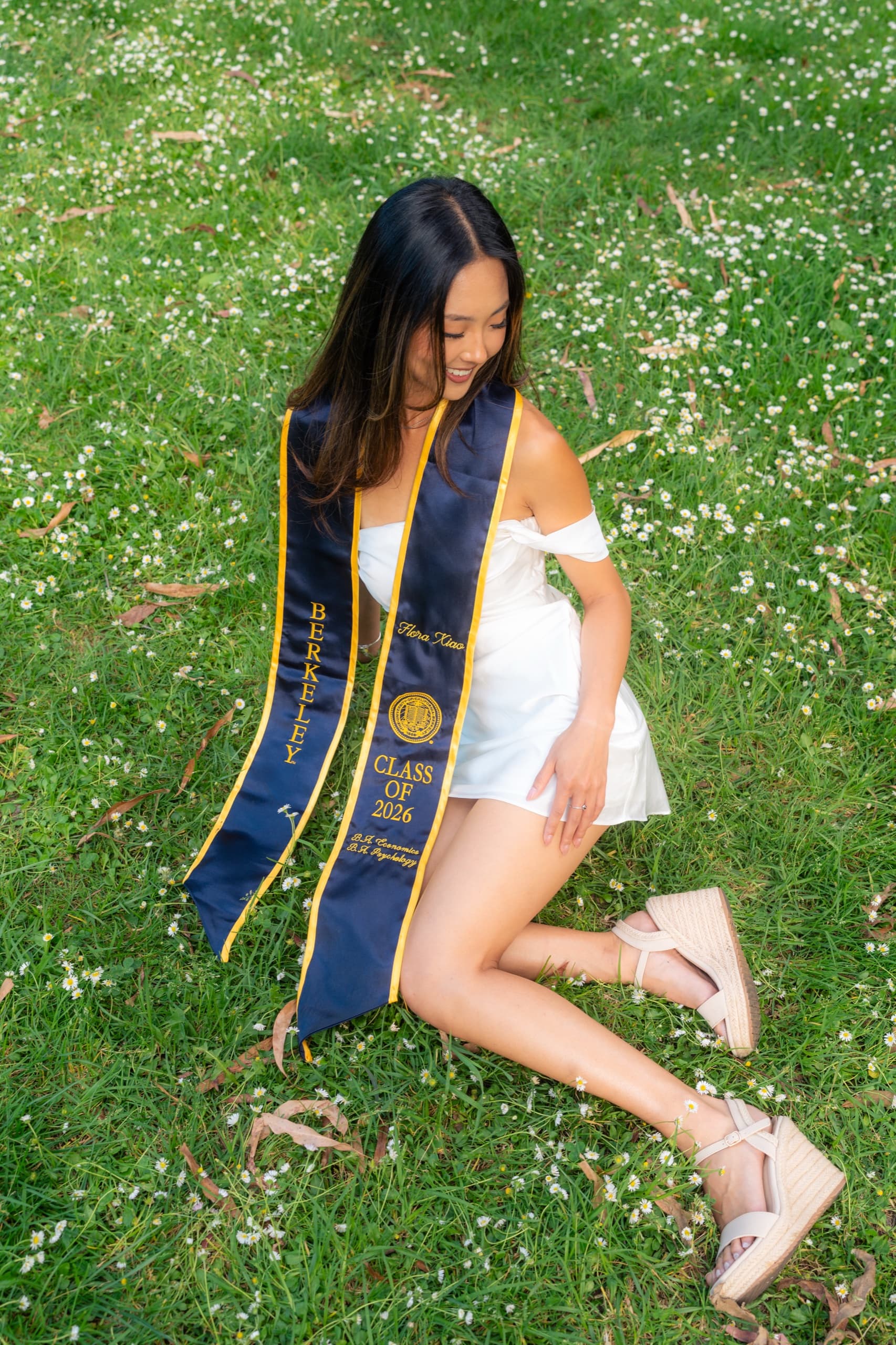 Overhead view of a graduate in a white off-shoulder dress and navy Berkeley Class of 2026 stole sitting on a lawn scattered with small white daisies, looking down and smiling.