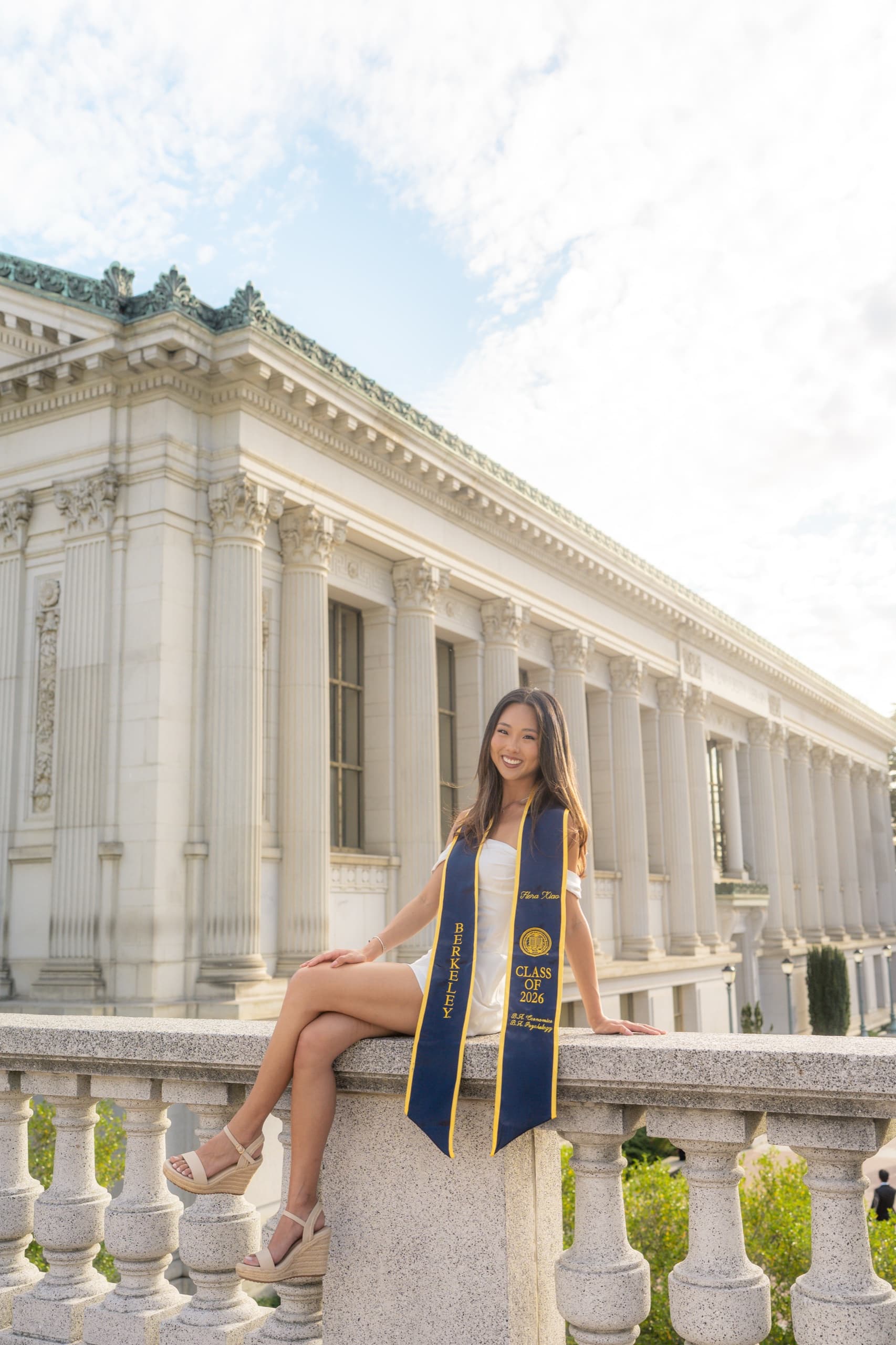 A graduate in a white dress and navy Berkeley Class of 2026 stole sits with her legs crossed on a stone balustrade in warm late-day light, with a large columned campus library and a cloudy sky behind her.
