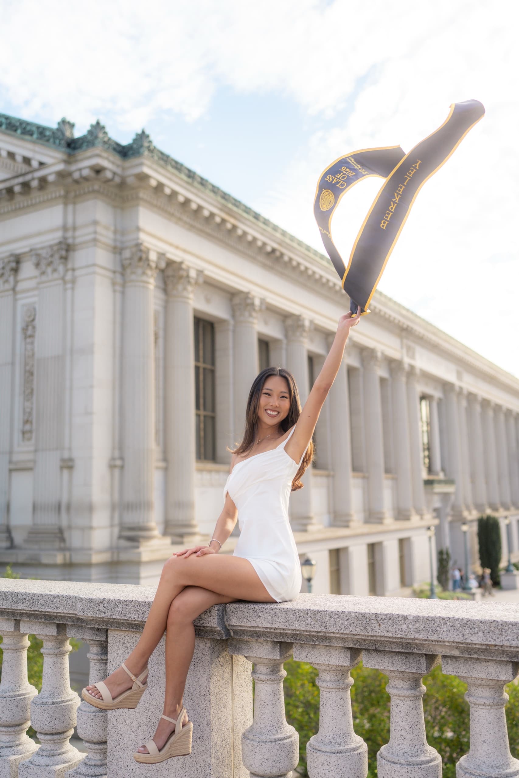 A graduate in a short white dress sits on a granite balustrade with one arm raised, holding a navy and gold Berkeley Class of 2026 stole that catches the wind, a columned neoclassical campus library behind her in bright afternoon light.