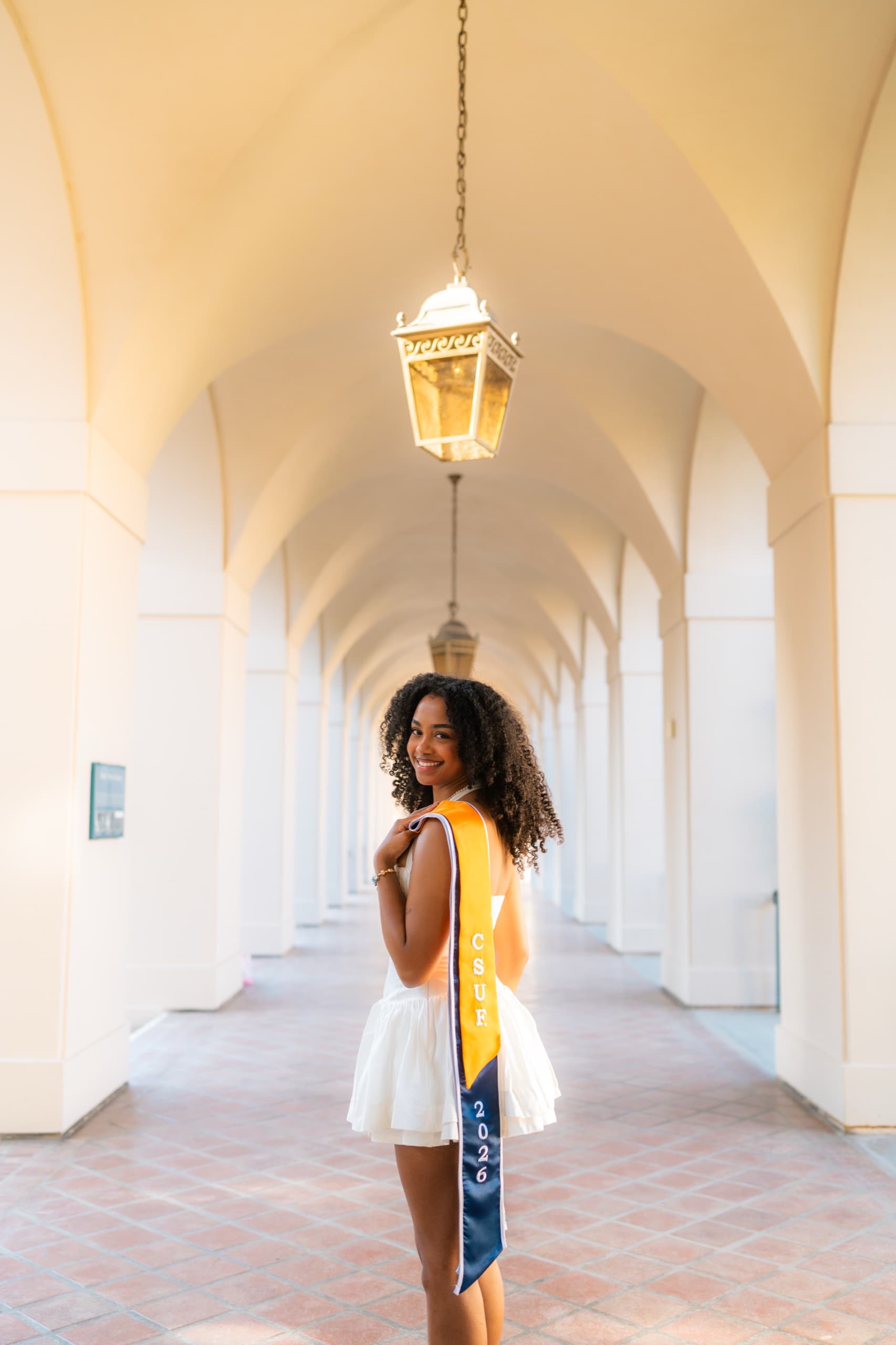 A graduate in a white dress with a gold and navy CSUF 2026 sash looks back over her shoulder while standing in a long white arched arcade with a terracotta tile floor and a lantern hanging above her.