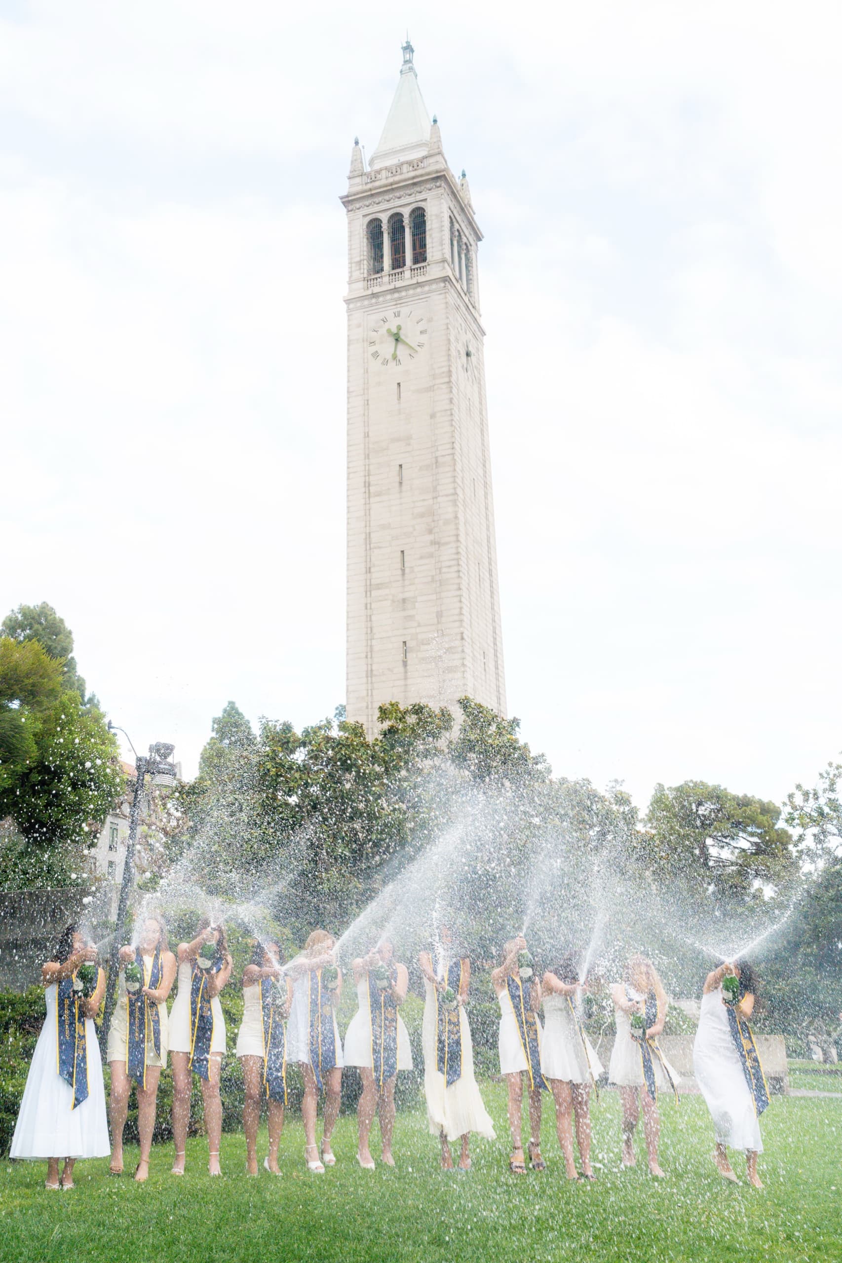 A row of about a dozen graduates in white dresses and navy Berkeley stoles stand on a lawn spraying champagne into the air in unison, with a tall stone campus clock tower rising behind them under an overcast sky.