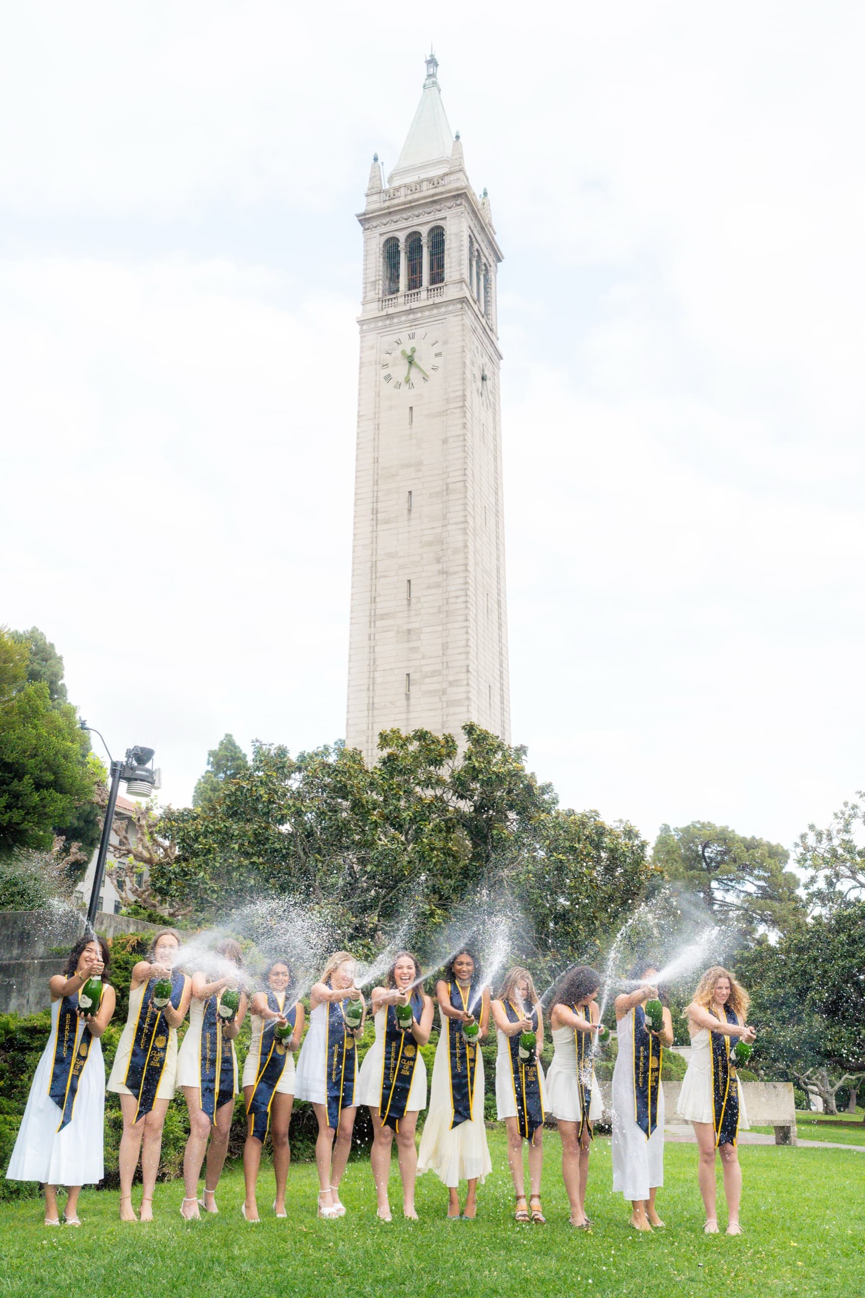 A long line of graduates in white dresses and navy and gold Berkeley Class of 2026 stoles spray champagne bottles across a green lawn, with a tall stone clock tower rising behind them under a bright overcast sky.