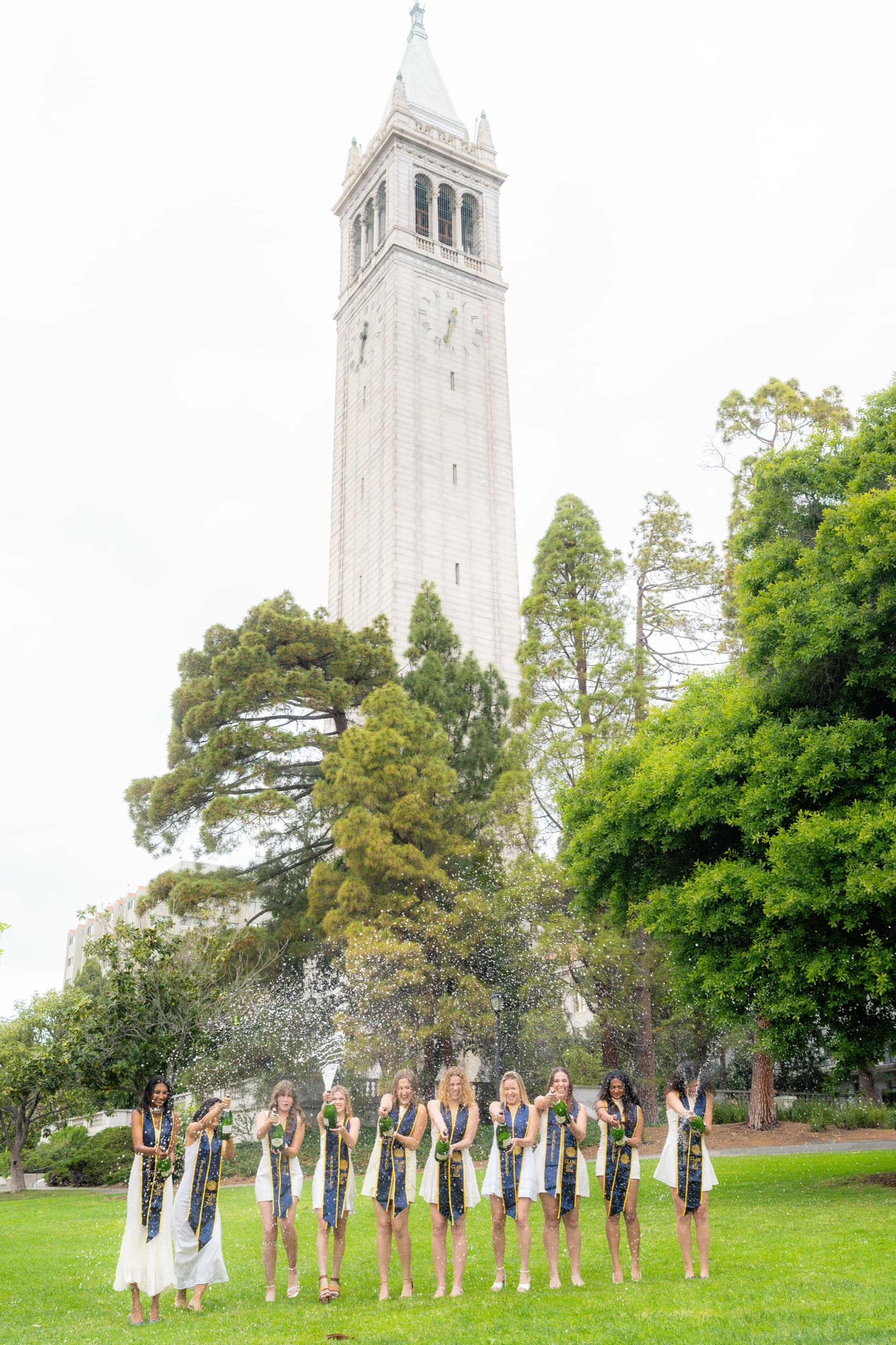 The same group of graduates in white dresses and navy Berkeley stoles stands spread across a lawn spraying champagne, photographed from farther back with tall trees framing the stone clock tower behind them.