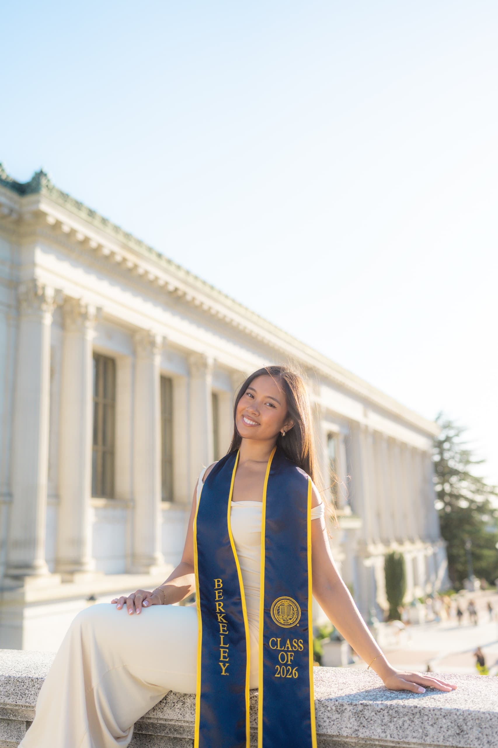 A graduate in a cream dress and a navy Berkeley Class of 2026 stole sits on a stone ledge with one hand resting beside her, in front of a columned neoclassical campus library, backlit by bright afternoon sun.