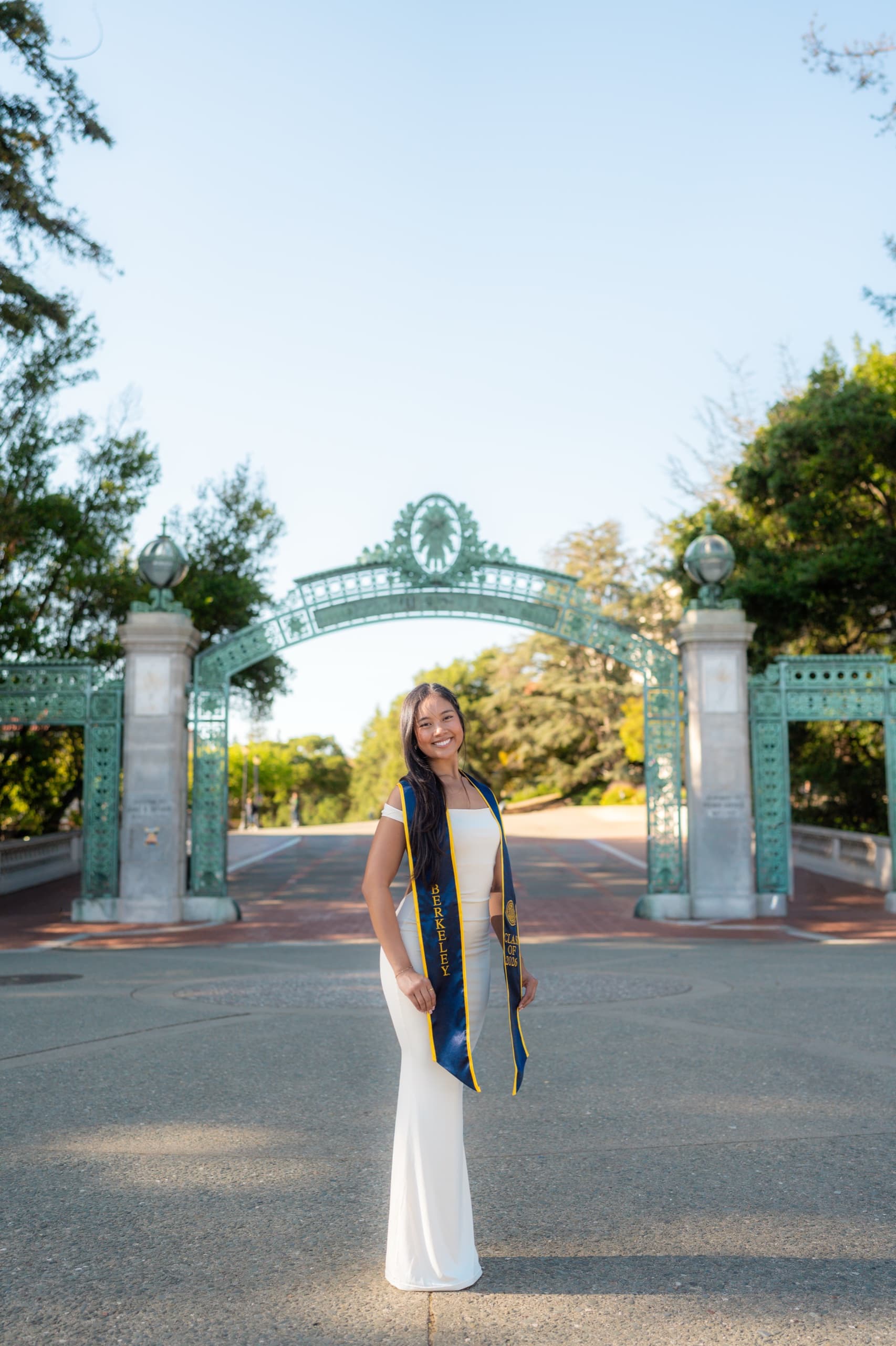 A graduate in a long white gown and a navy Berkeley Class of 2026 stole stands centered on an empty paved walkway in front of an ornate green metal campus gate, lit by low late-afternoon sun under a clear blue sky.