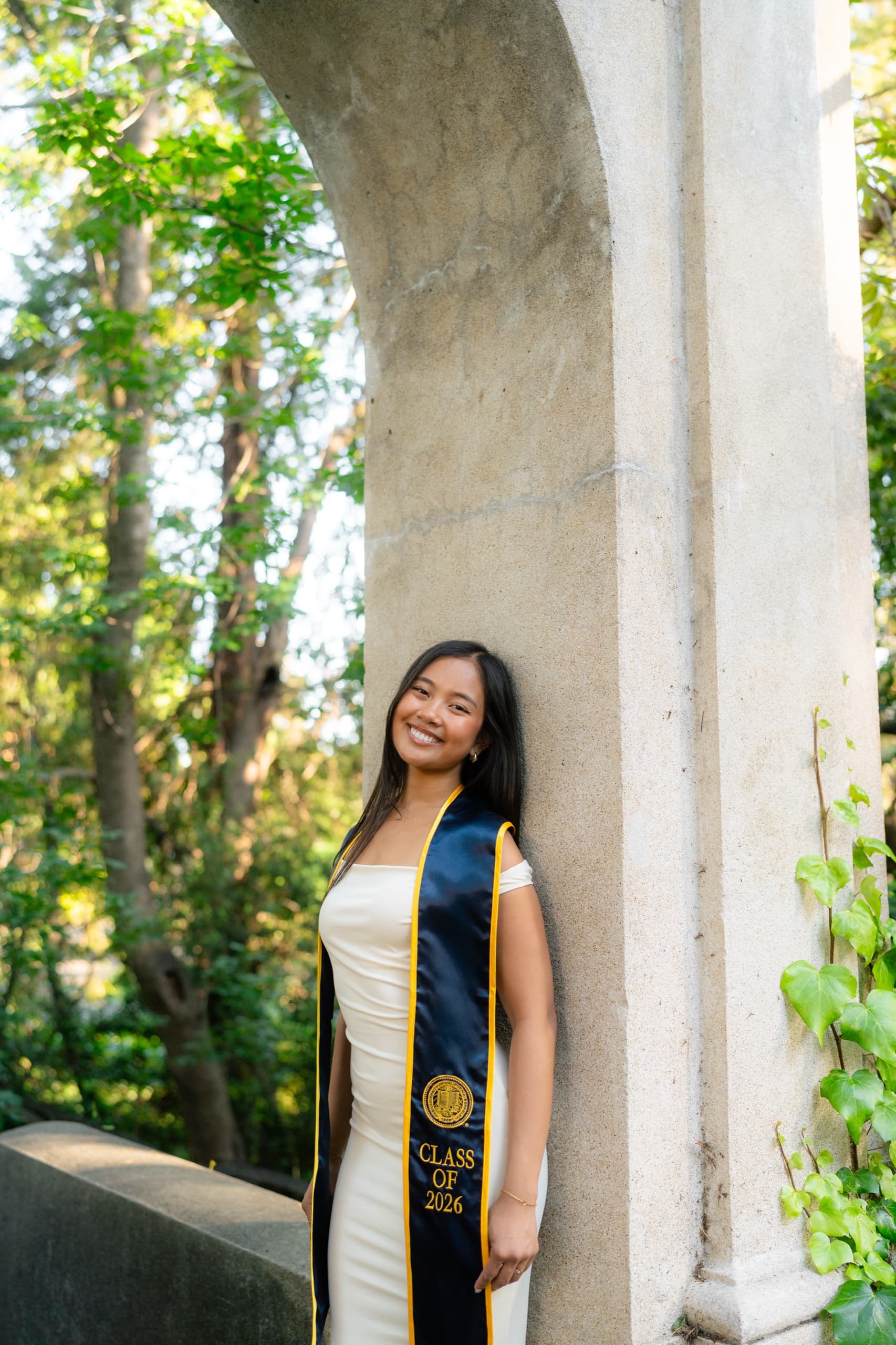 A graduate in an ivory gown and a navy Berkeley Class of 2026 stole leans against a weathered concrete arch column, smiling toward the camera, with sunlit green trees and climbing ivy behind her.