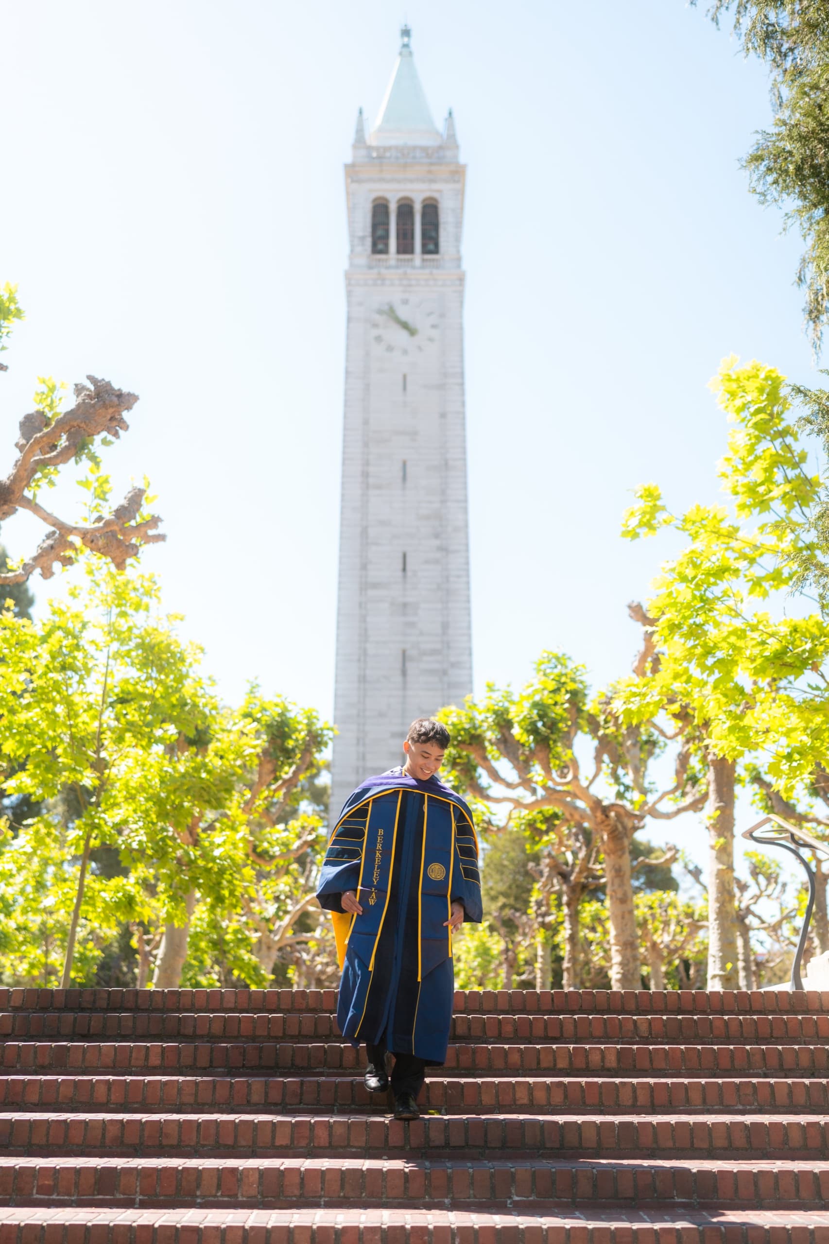 A graduate in navy and gold doctoral regalia walks down wide brick steps, looking down at his feet, with the tall white clock tower of the Campanile rising behind him between bright green spring trees.
