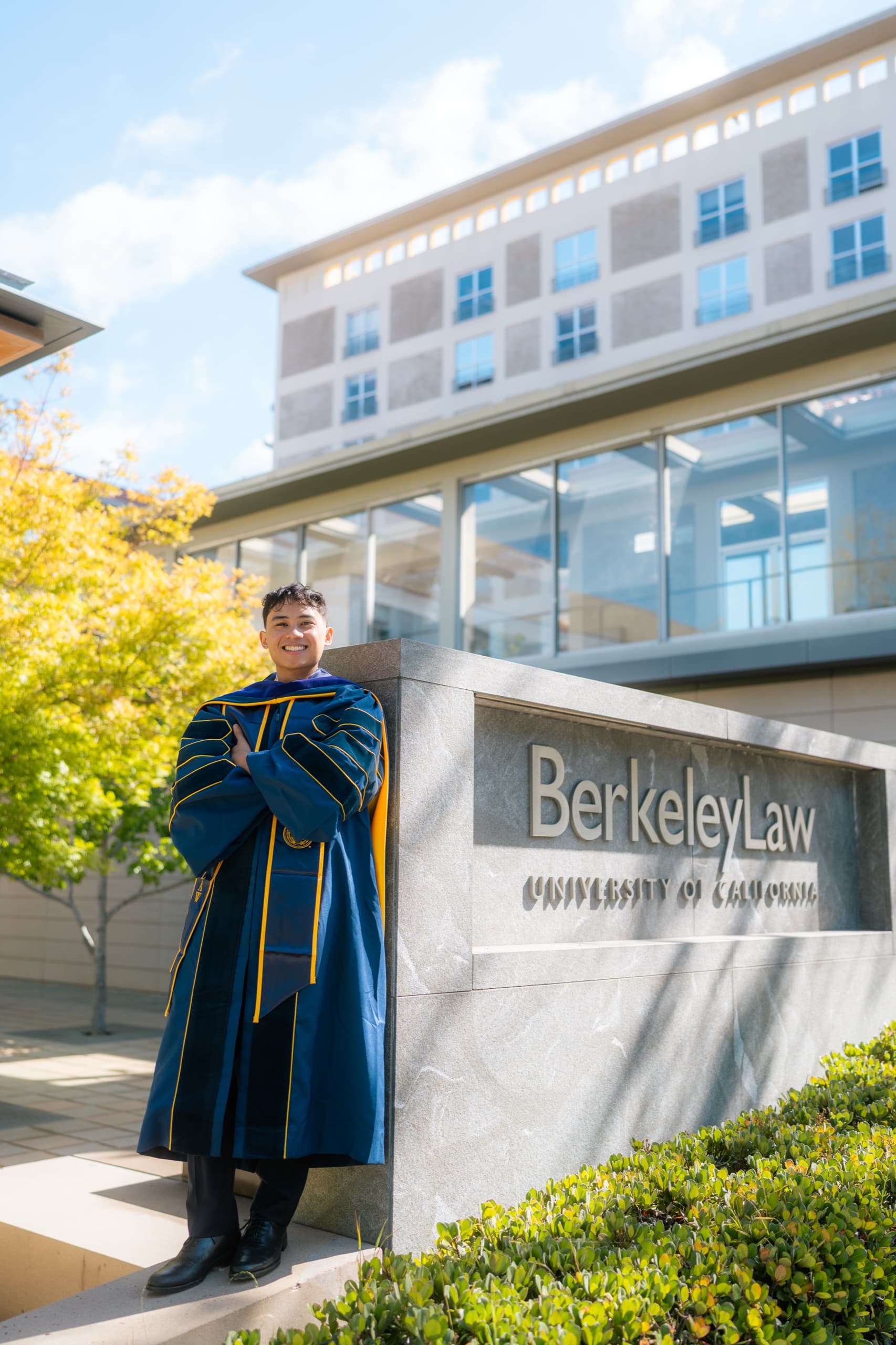 A graduate in navy and gold doctoral regalia stands with arms folded, leaning against a granite sign reading Berkeley Law, University of California, with a glass-walled campus building and bright midday sky behind him.