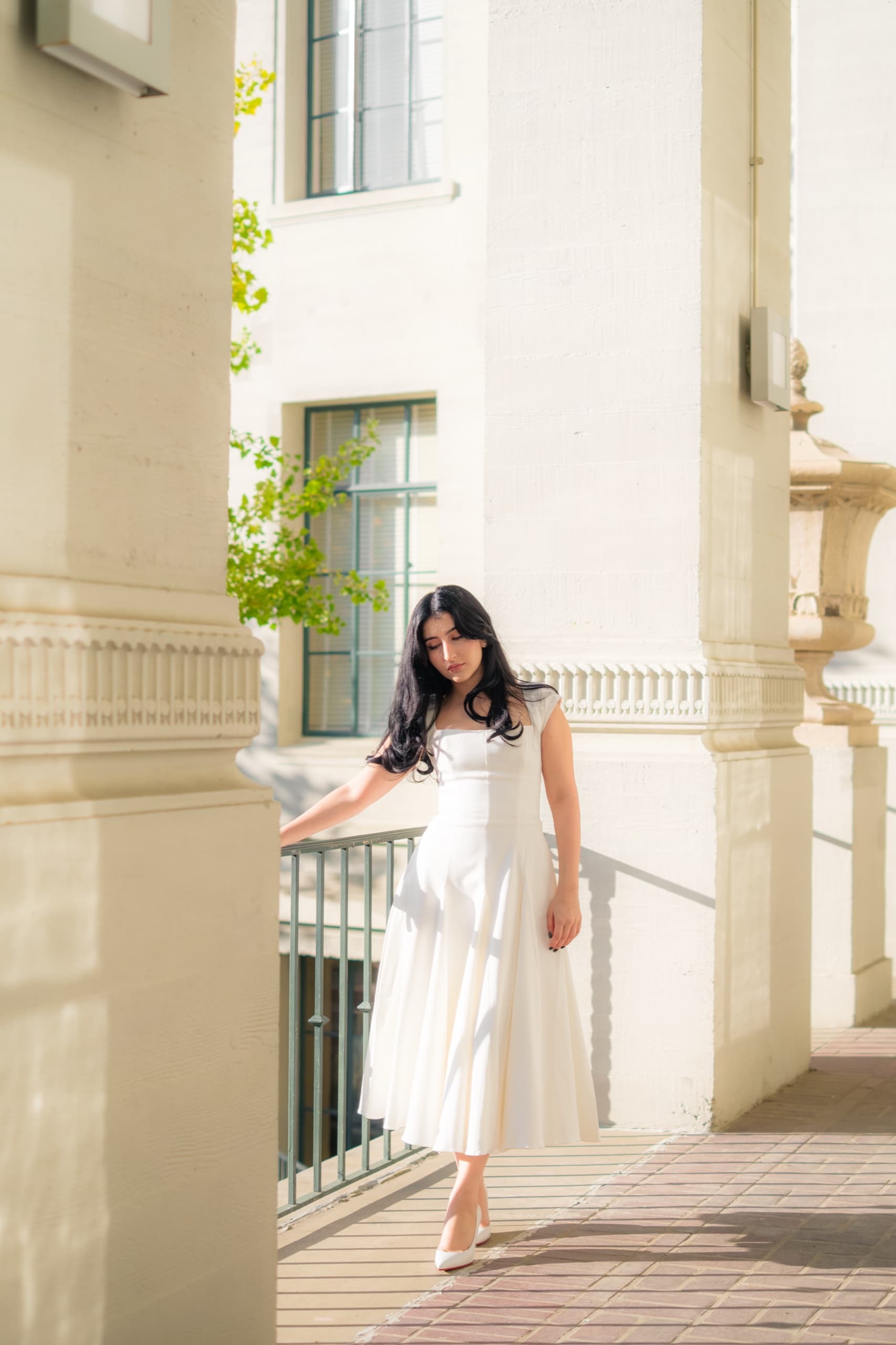 A graduate in a white cap-sleeve midi dress and white heels stands on a brick walkway beside a green metal railing, one hand resting on the rail and her gaze lowered, framed by the pale columns and balustrade of a classical campus building in warm sunlight.