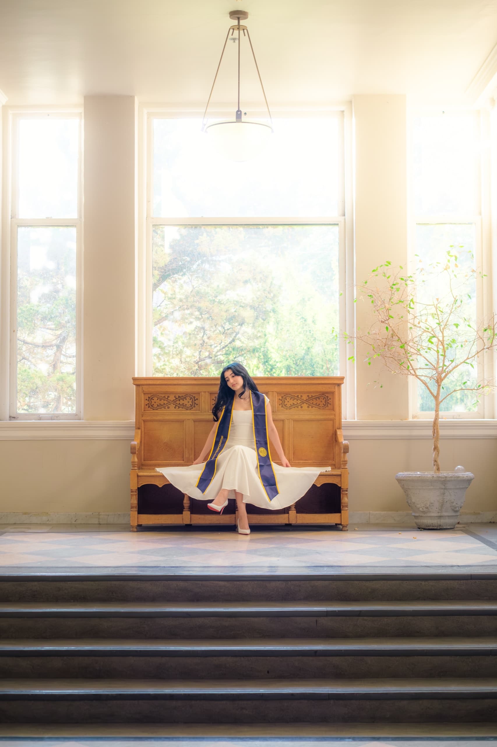 A graduate in a white dress and a navy Berkeley stole sits on a carved oak bench in a bright interior hall, holding her skirt spread wide across the seat, with tall windows and a potted tree behind her and a checkered marble floor and steps in the foreground.