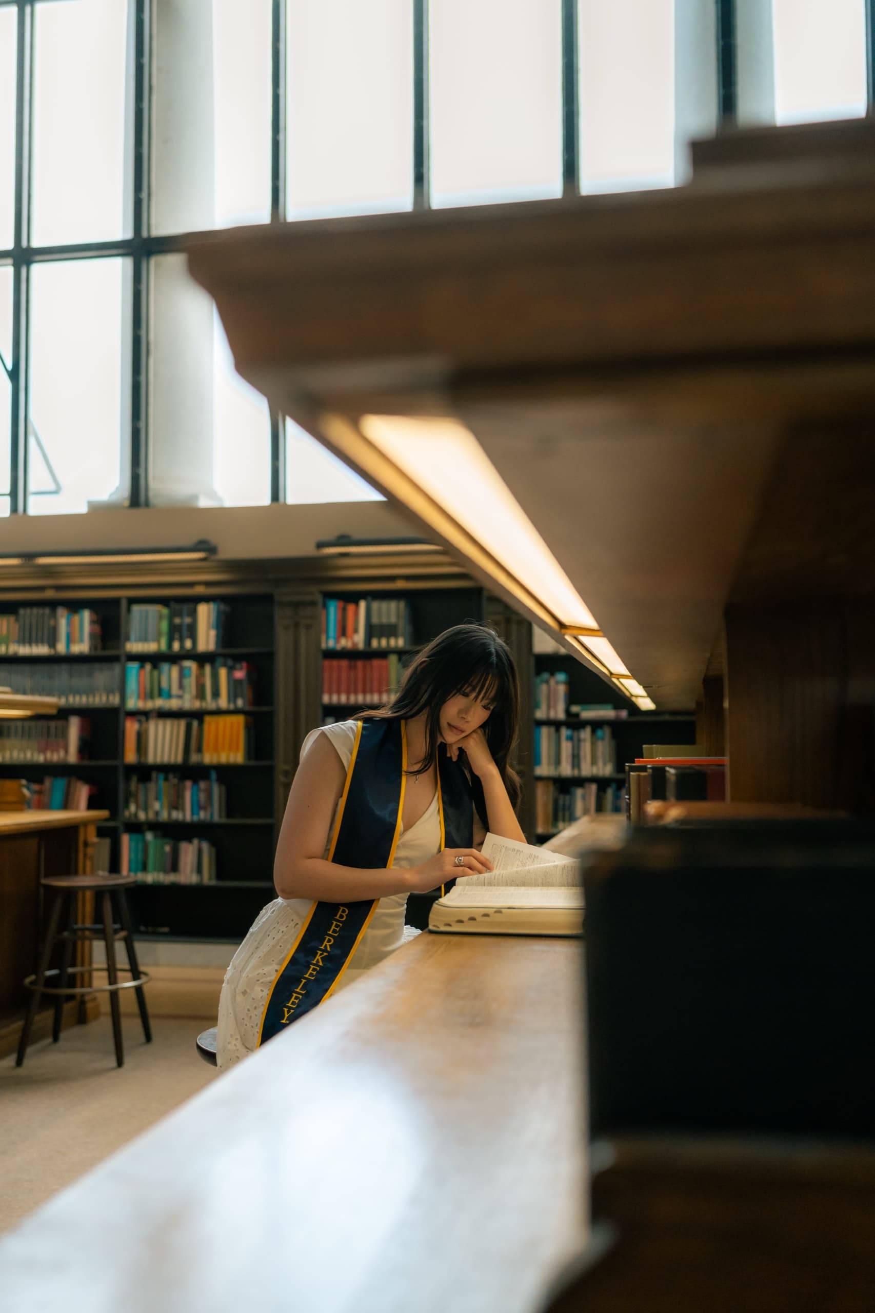 A graduate in a white dress and navy Berkeley stole leans on a long wooden library table, reading an open book beneath tall windows with tiers of bookshelves behind her.
