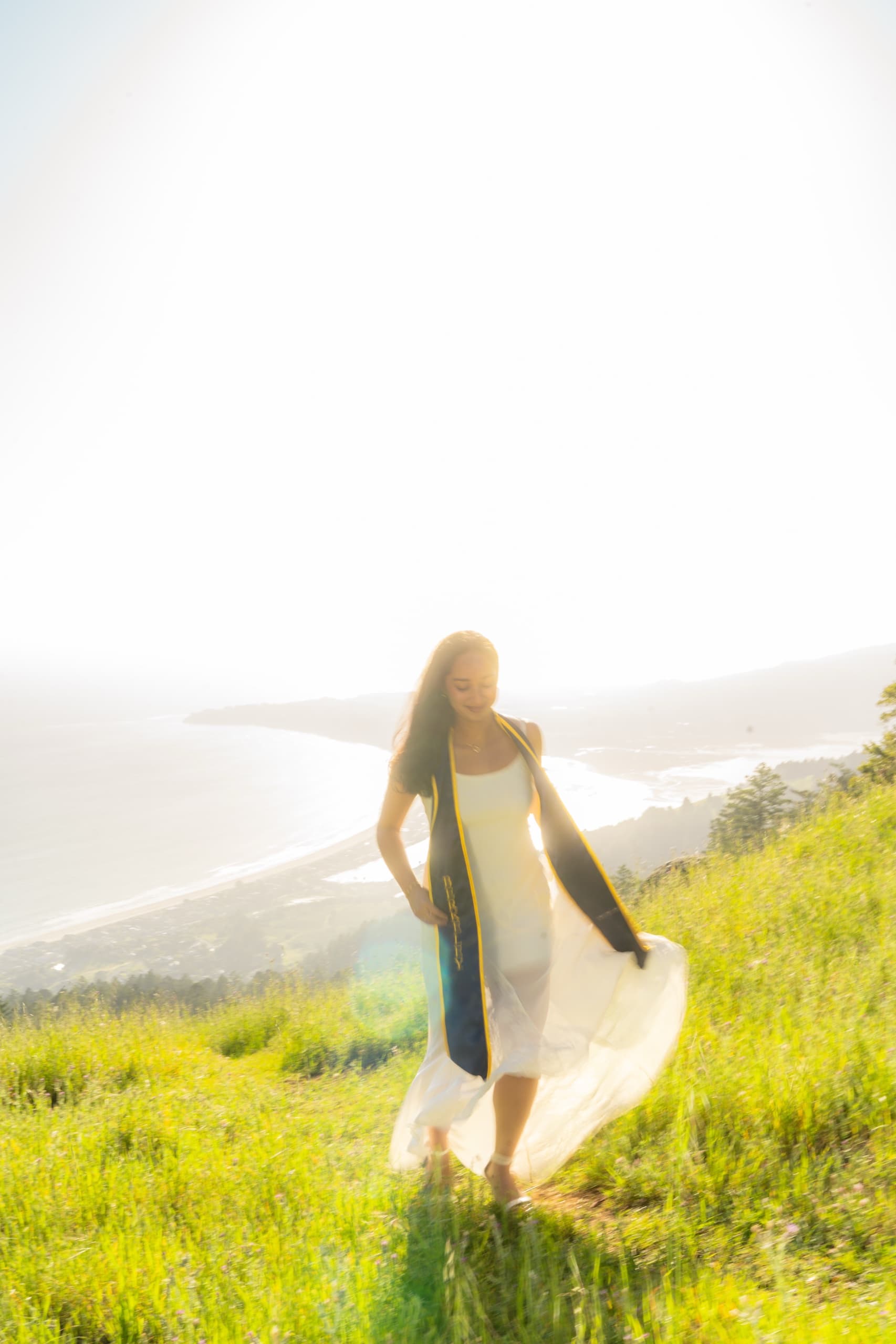 A graduate in a white dress and navy stole walks down a green grass hillside into strong backlight, with a hazy coastline, lagoon, and ocean spread out far below her.