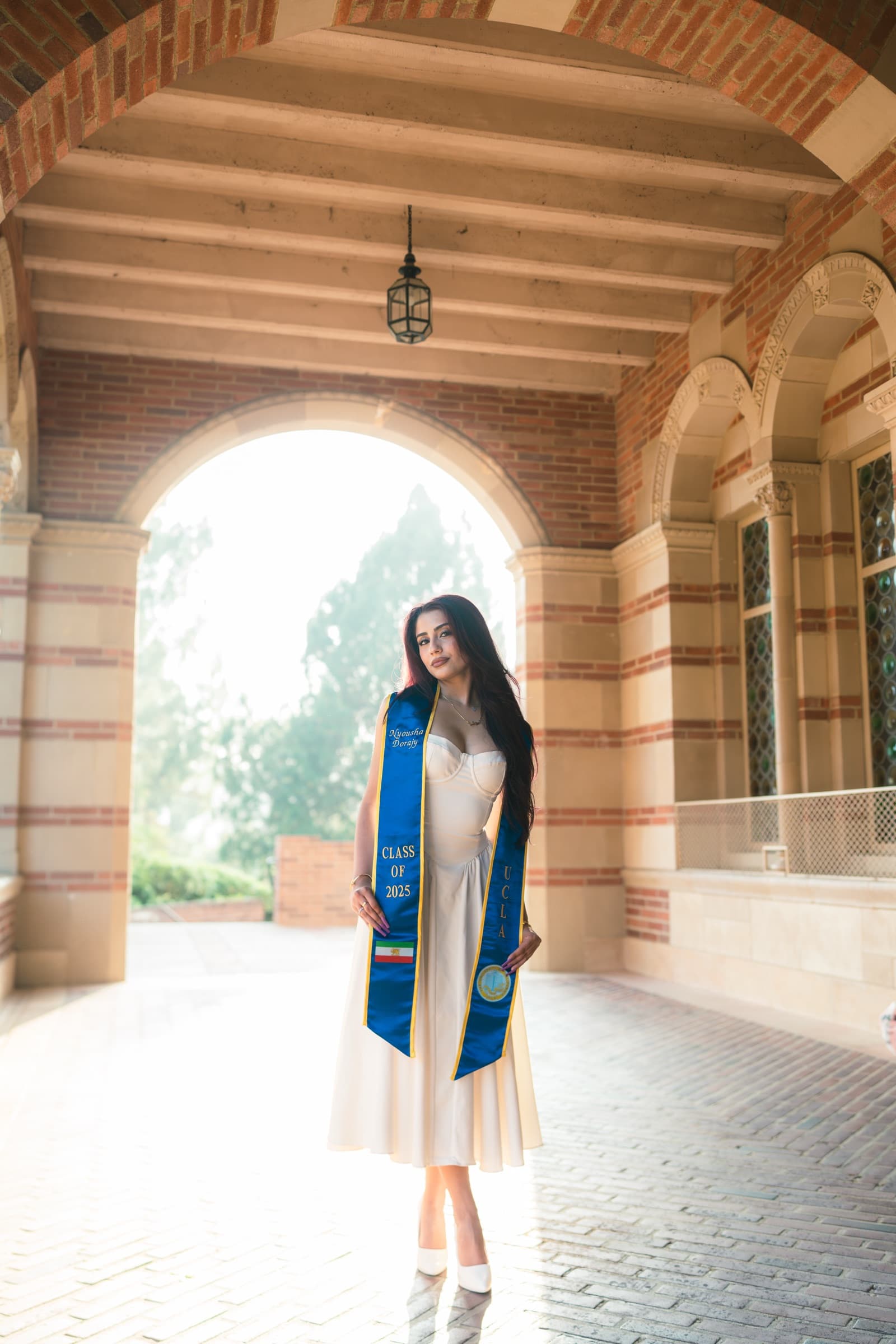 Graduate in a white dress and Class of 2025 stole framed by a brick archway colonnade