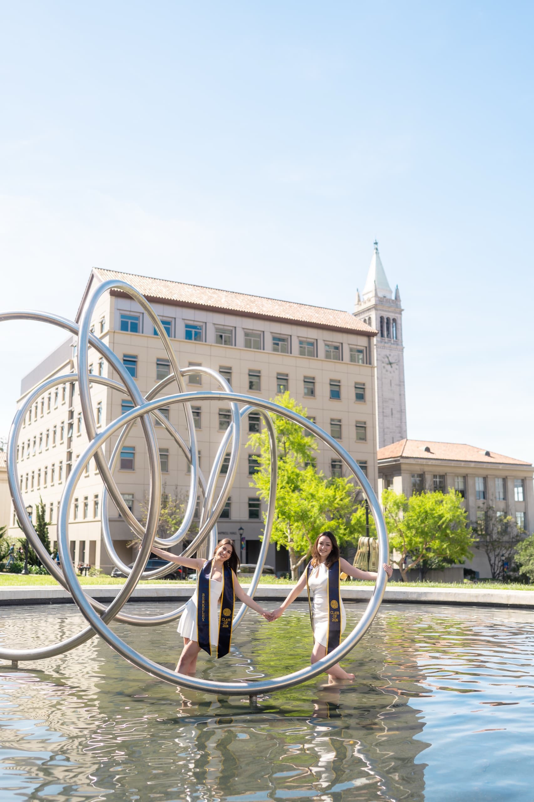 Two graduates in white dresses and navy Class of 2026 stoles stand barefoot in a shallow campus fountain, holding hands inside a large chrome loop sculpture, with a clock tower and campus buildings behind them under a clear sky.