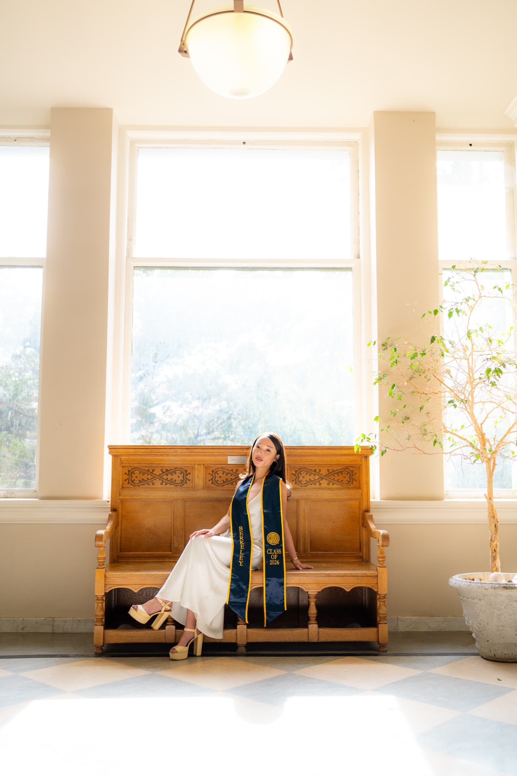 A graduate in a white dress and navy Class of 2026 stole sits on a carved wooden bench in a bright hall, centered between three tall windows, with a checkered tile floor and a potted tree beside her.