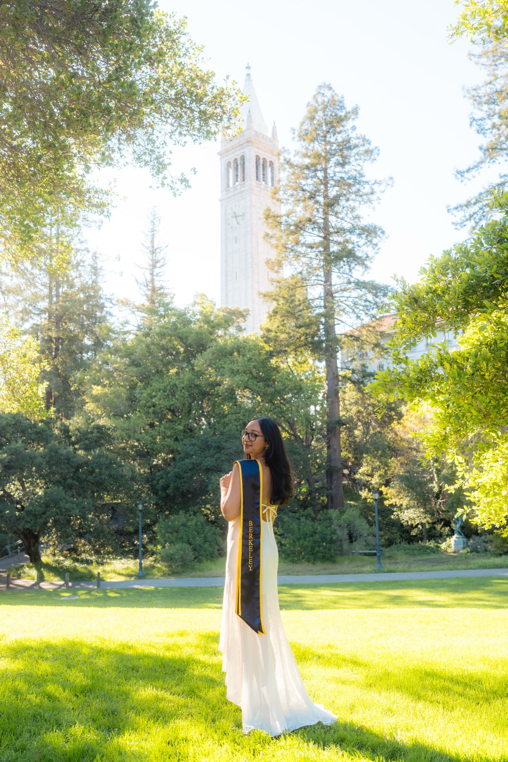A graduate in a long cream dress with a navy and gold Berkeley stole stands on a sunlit lawn and looks back over her shoulder, with a tall white campus bell tower rising through the trees behind her.