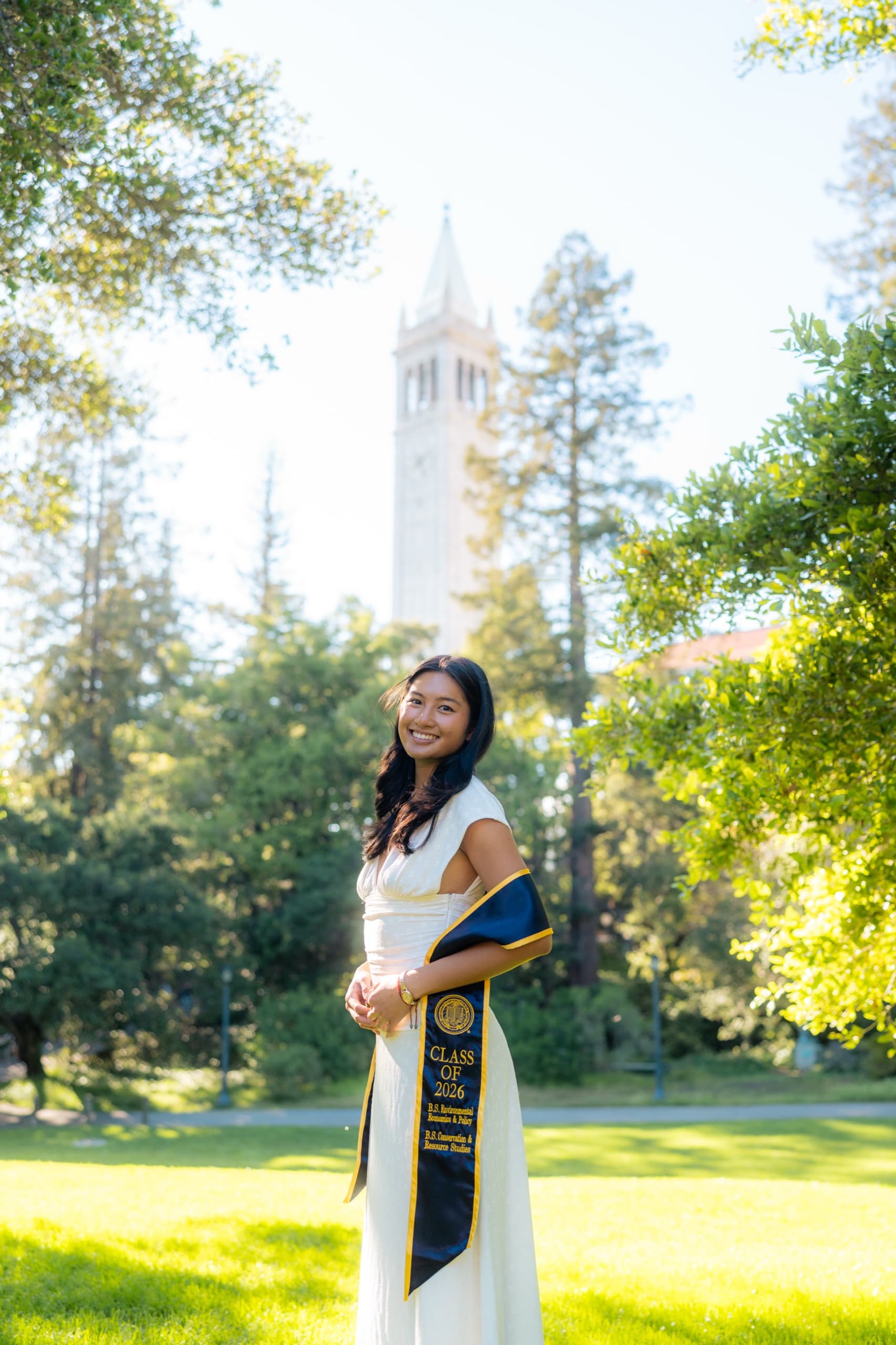 A graduate in a cream dress smiles toward the camera with a navy Class of 2026 Berkeley stole draped over one arm, standing on a lawn in late afternoon sun with a tall white campus bell tower in the distance behind her.
