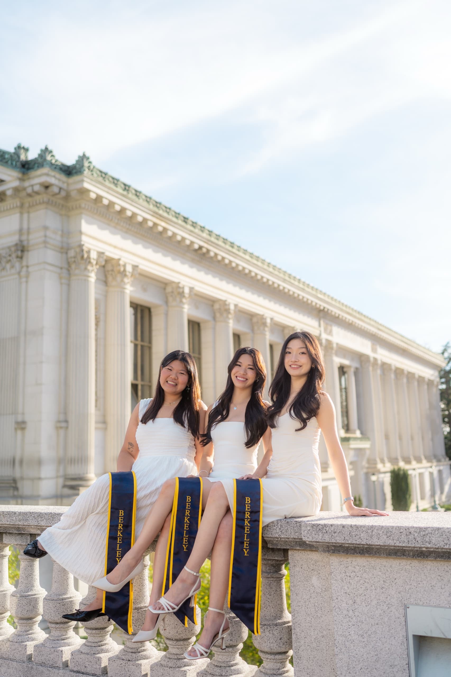 Three graduates in white dresses sit side by side on a stone balustrade with their navy and gold Berkeley stoles hanging over the edge, in front of a neoclassical campus library with a long row of columns.