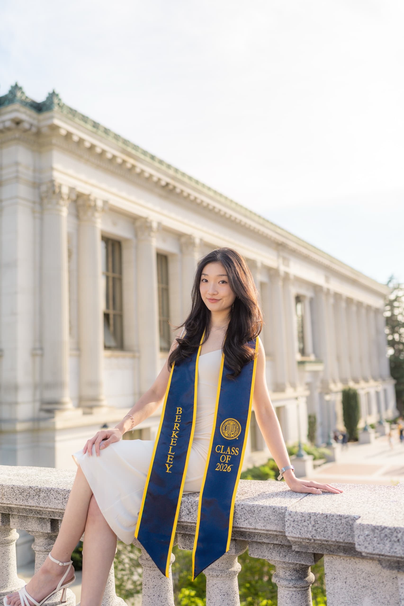 A graduate in a cream dress sits on a stone balustrade wearing a navy and gold Berkeley Class of 2026 stole, one hand resting on the ledge behind her, with a columned campus library receding into soft afternoon light.