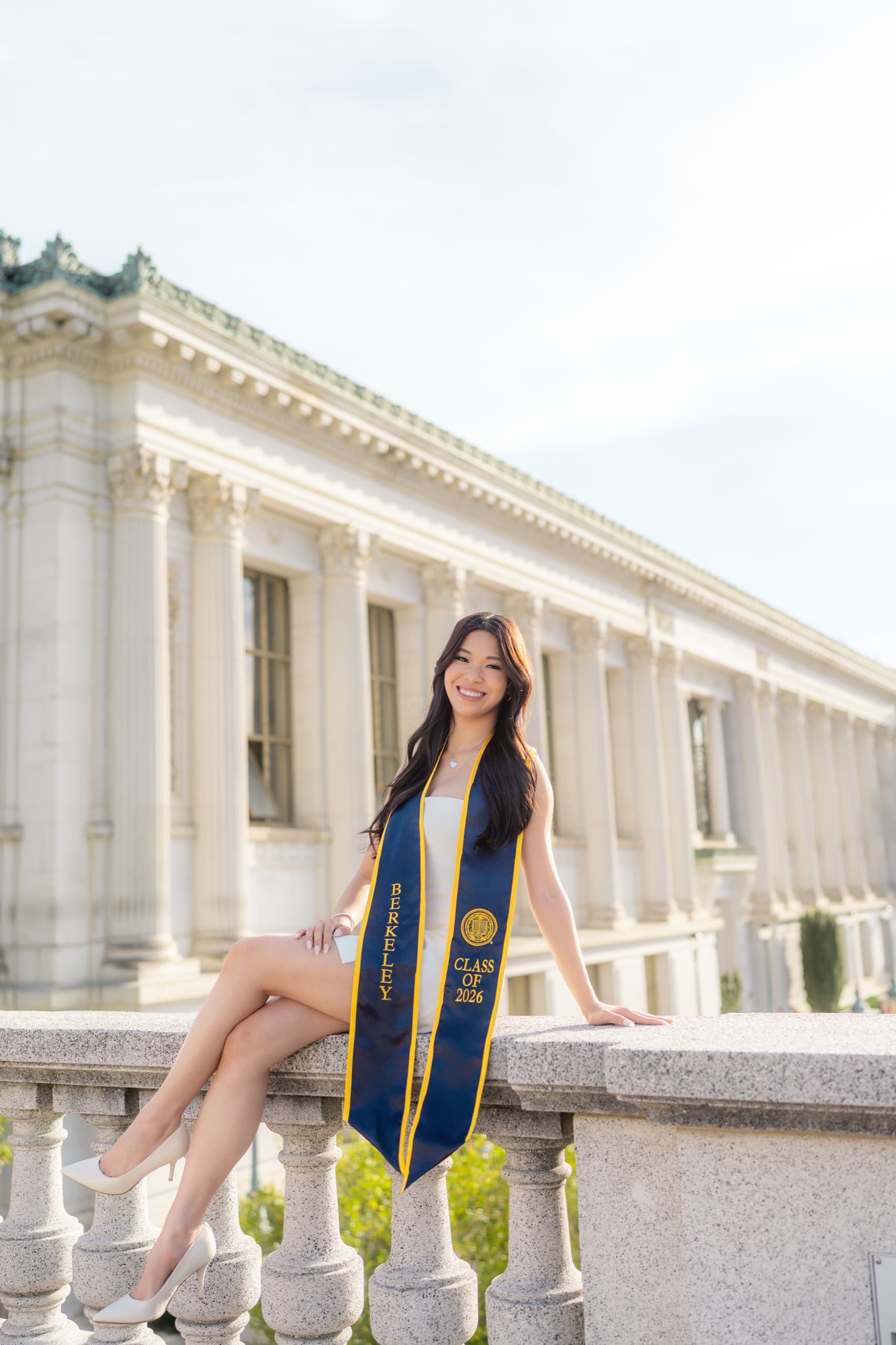A graduate in a short white dress sits on a stone balustrade with her legs crossed and a navy Berkeley Class of 2026 stole around her neck, smiling toward the camera in front of a long columned library facade.