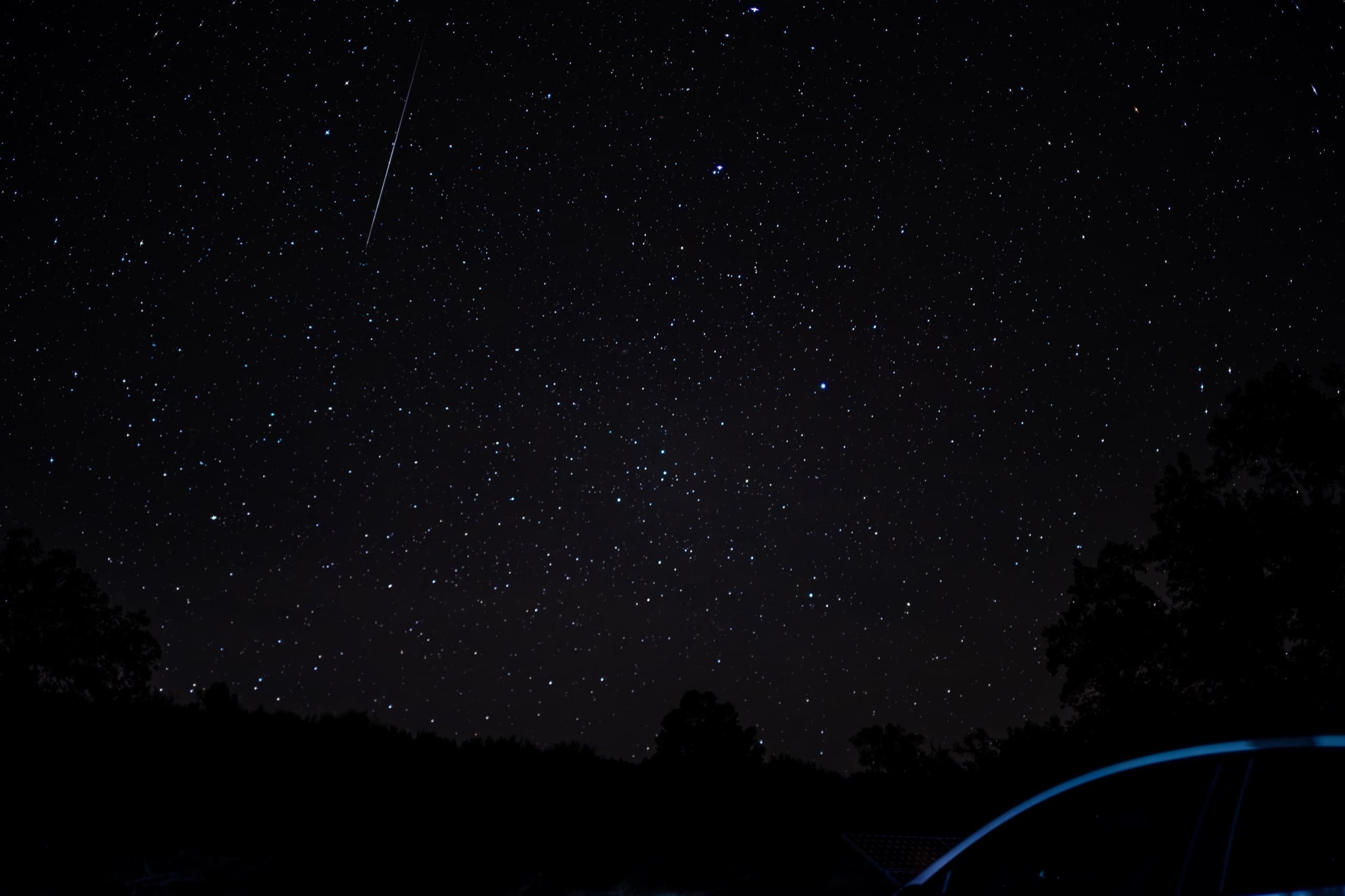 A long, bright meteor streaks above the tree line while the roofline of a parked car catches faint blue light at the corner of the frame.