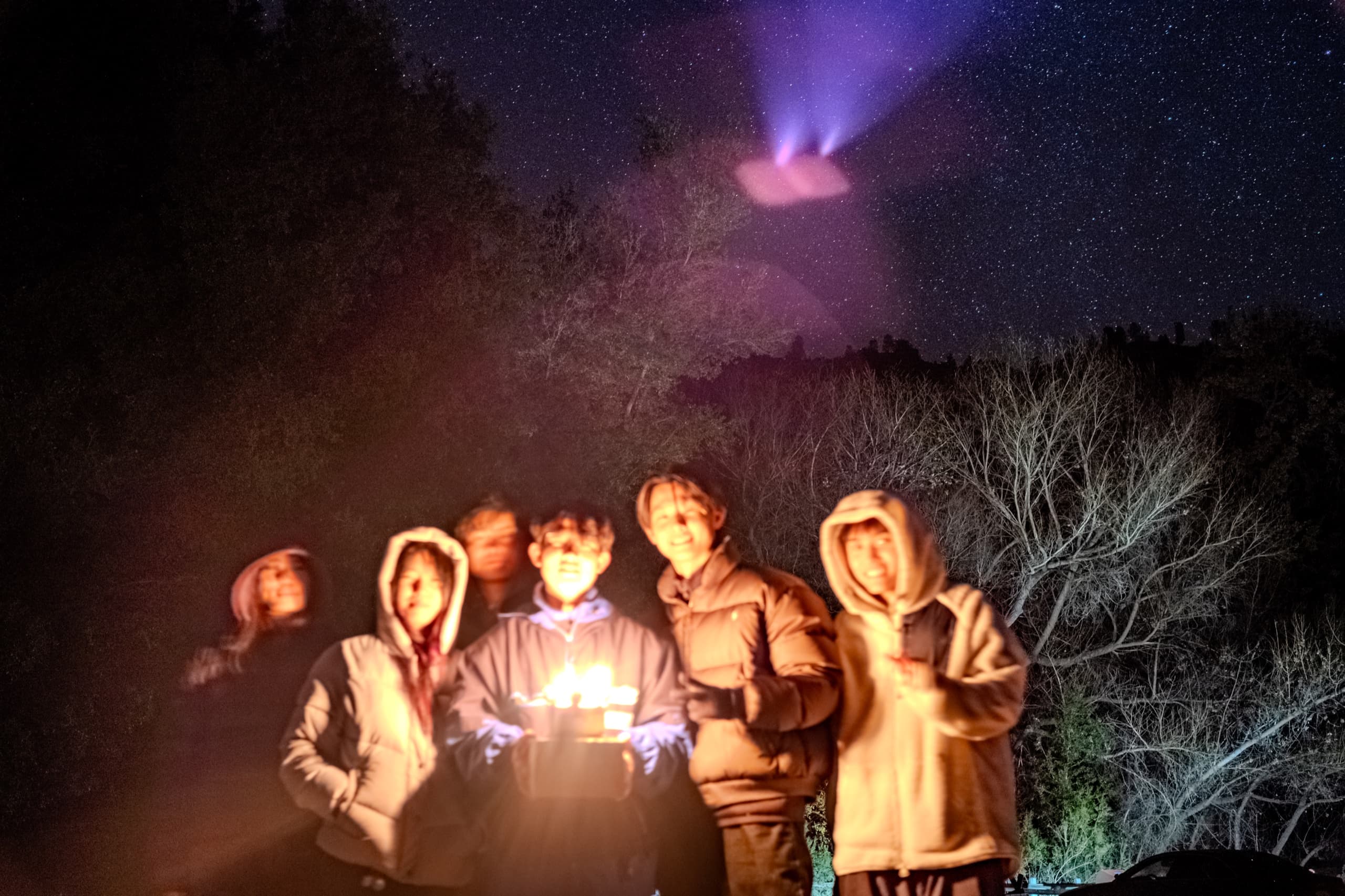 Six friends in winter jackets crowd around lit birthday candles under a starry sky, faces warm and motion-blurred in the eight-second exposure.