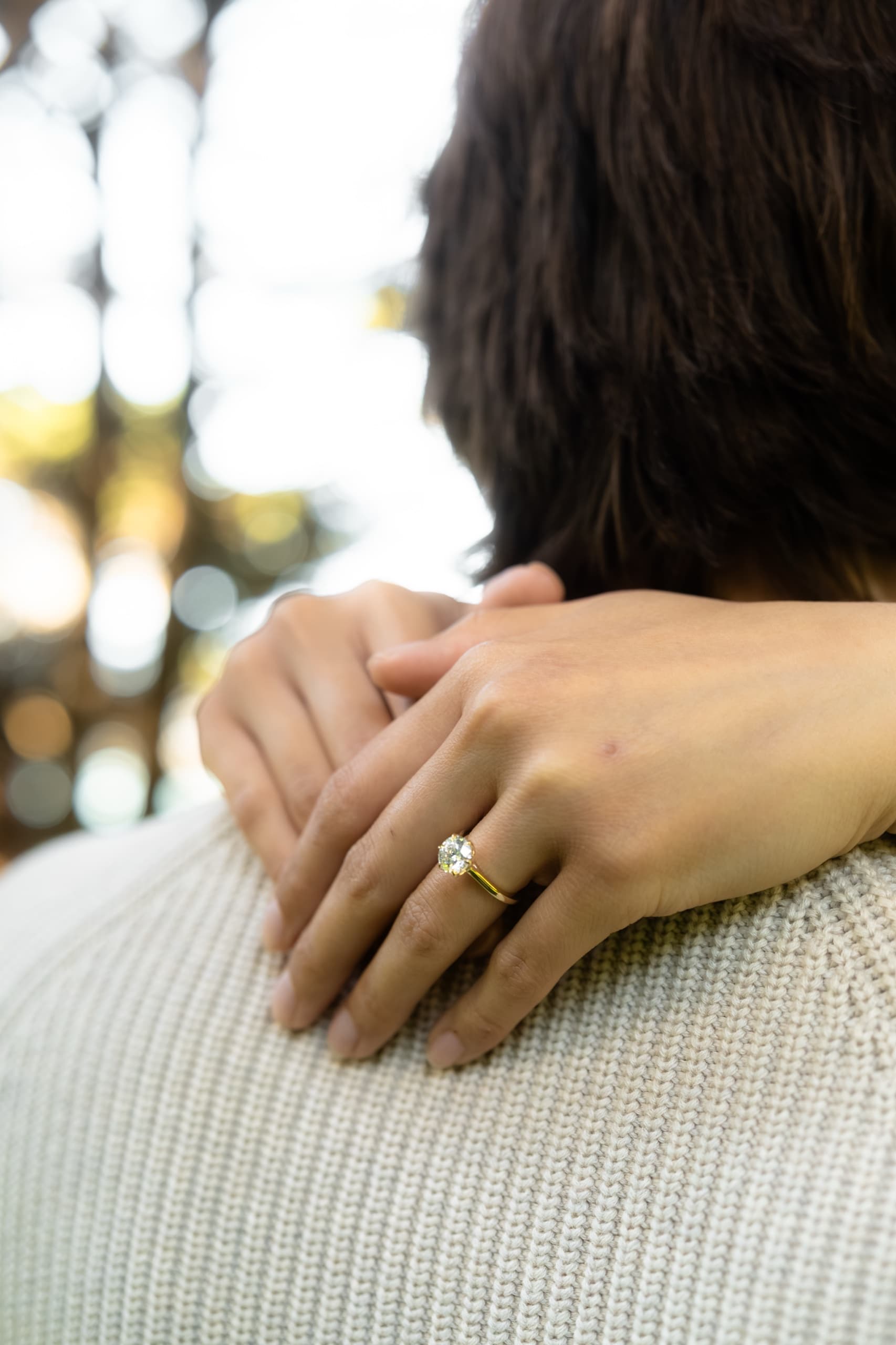 A woman's hands resting on her partner's shoulder after a proposal, an oval engagement ring on her finger, backlit trees blurred behind them.