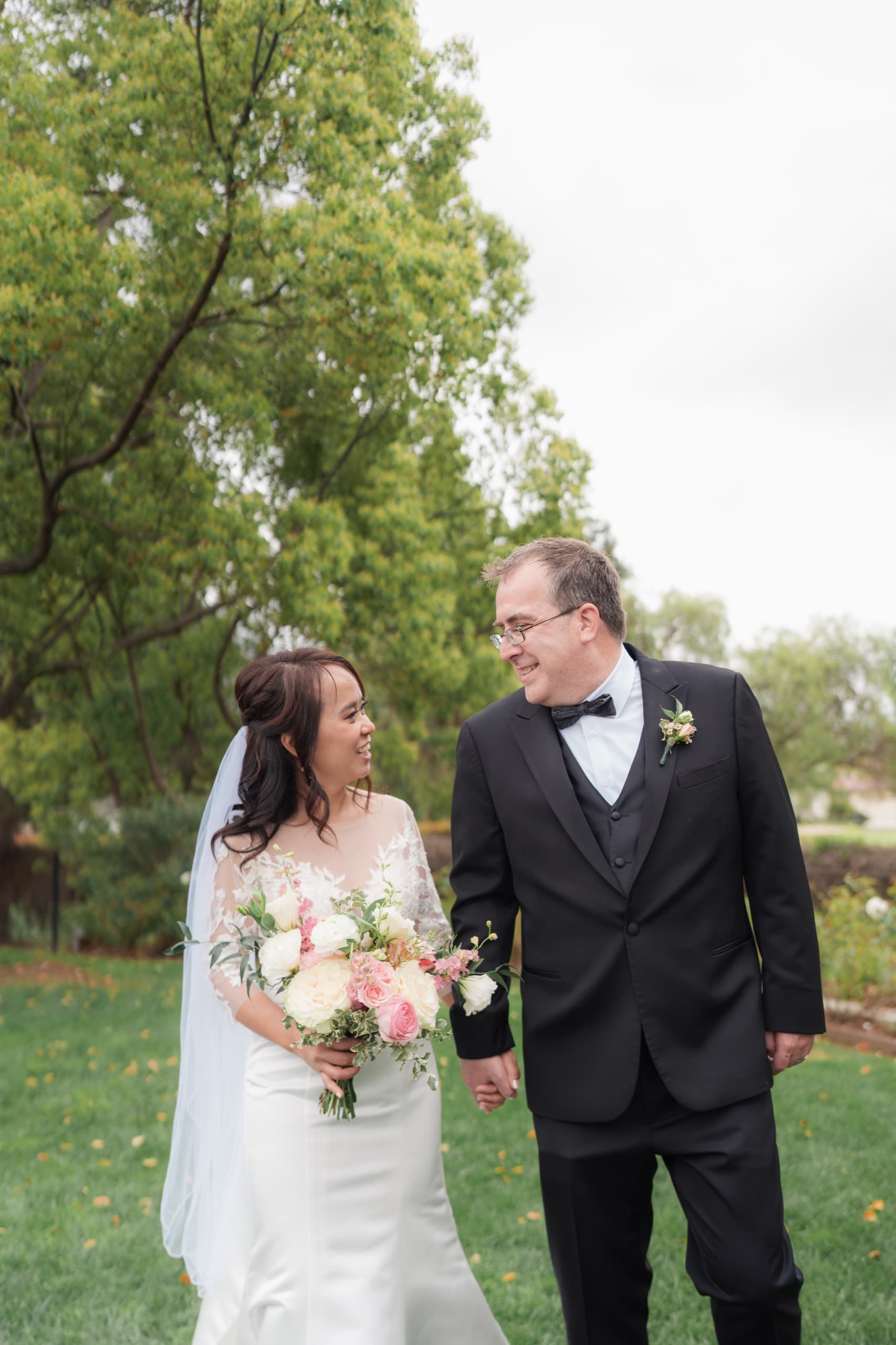 A bride and groom walking hand in hand across a garden lawn.