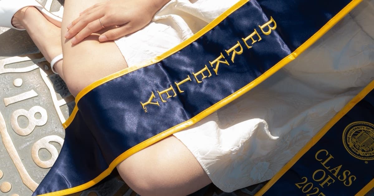 A navy and gold graduation stole against a white dress and bronze university seal