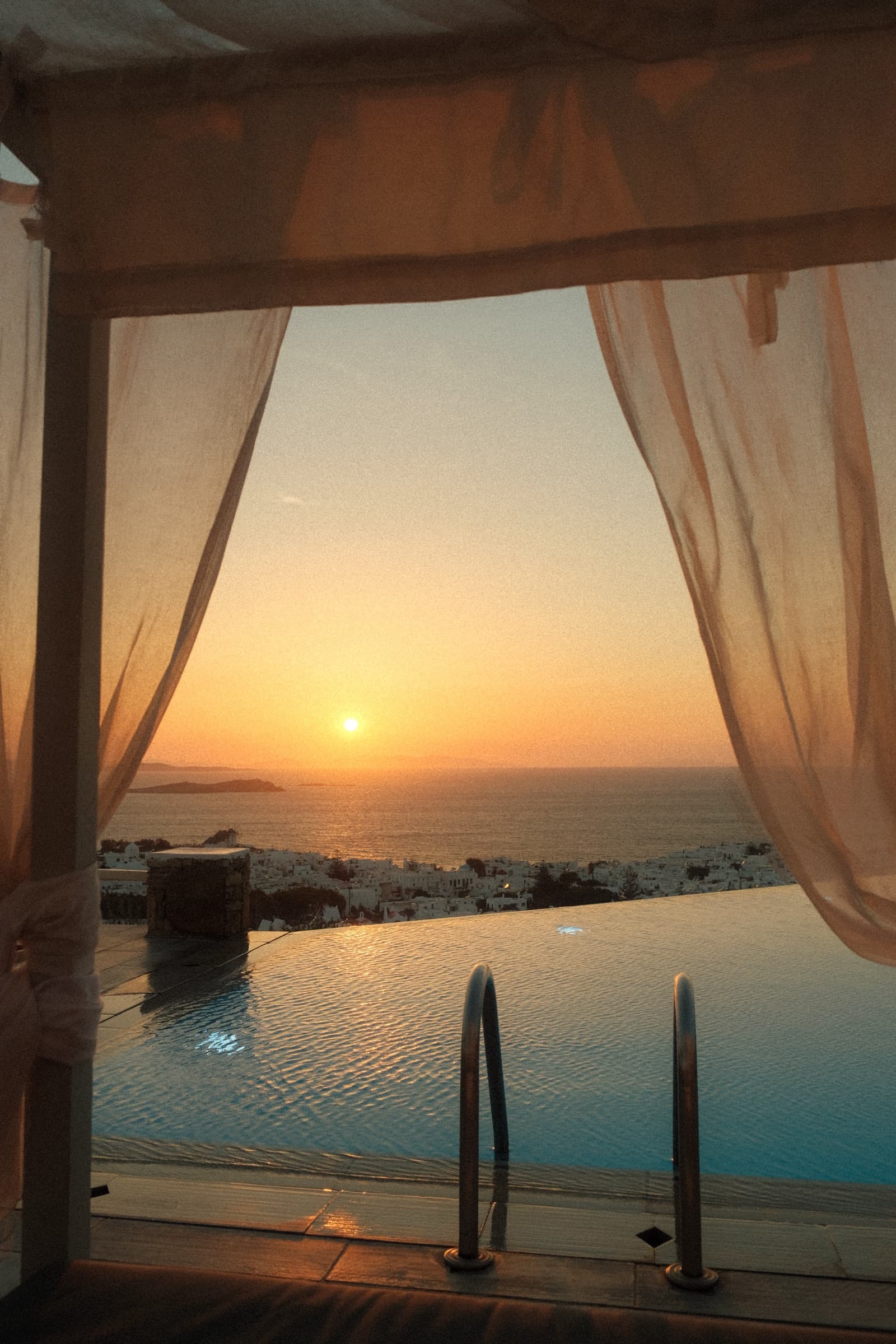 Sunset over the Aegean from a cabana, sheer curtains framing an infinity pool