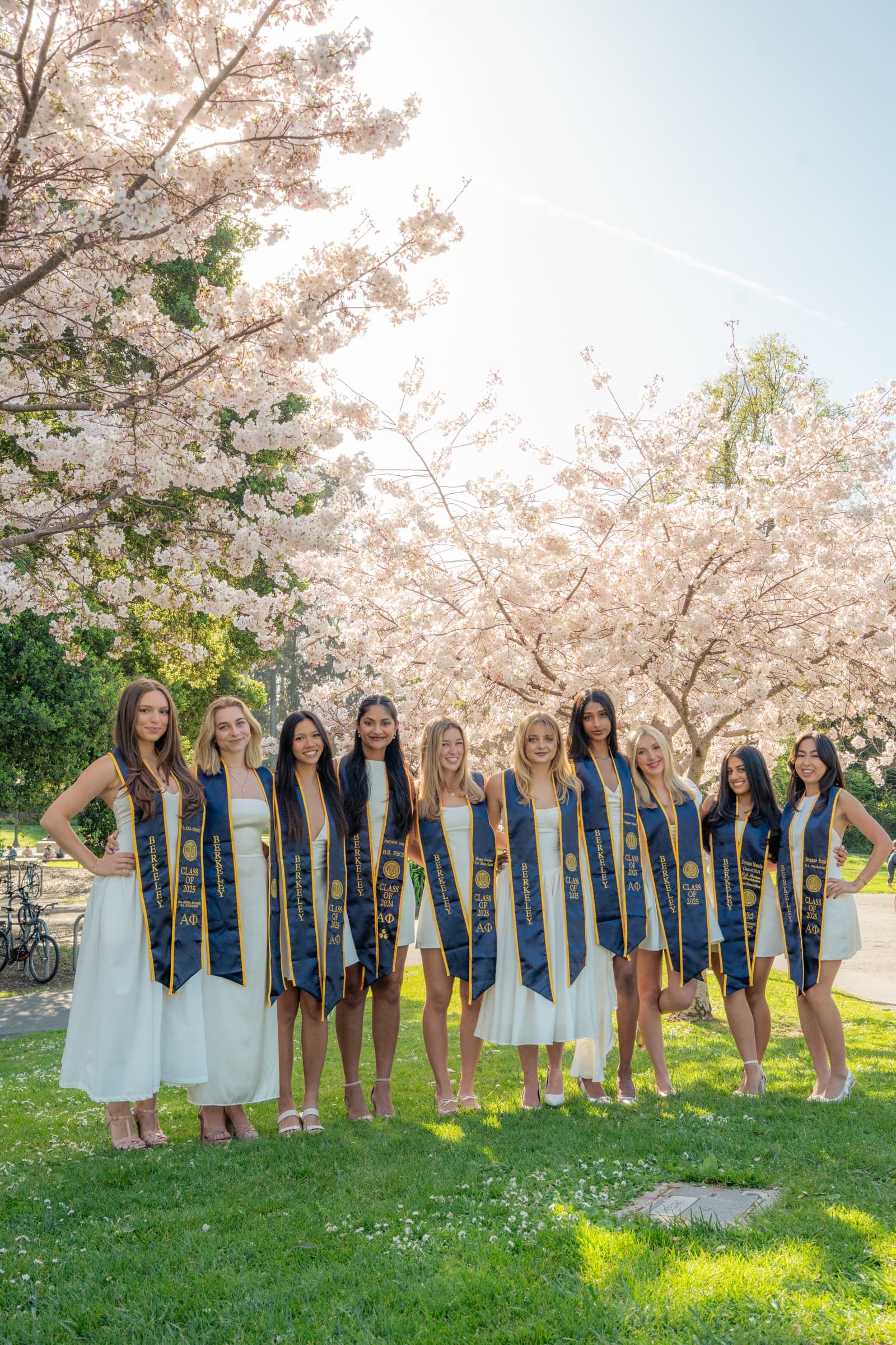 Ten graduates in white dresses and matching stoles lined up under blooming cherry trees