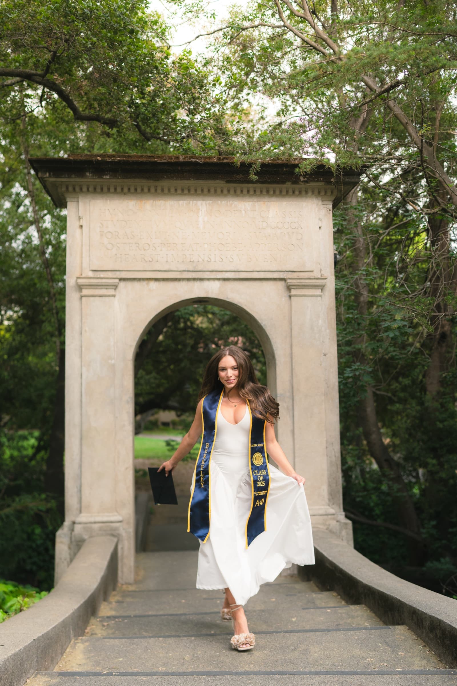 Graduate in a white dress and Cal stole mid-stride beneath a stone arch on a campus footbridge