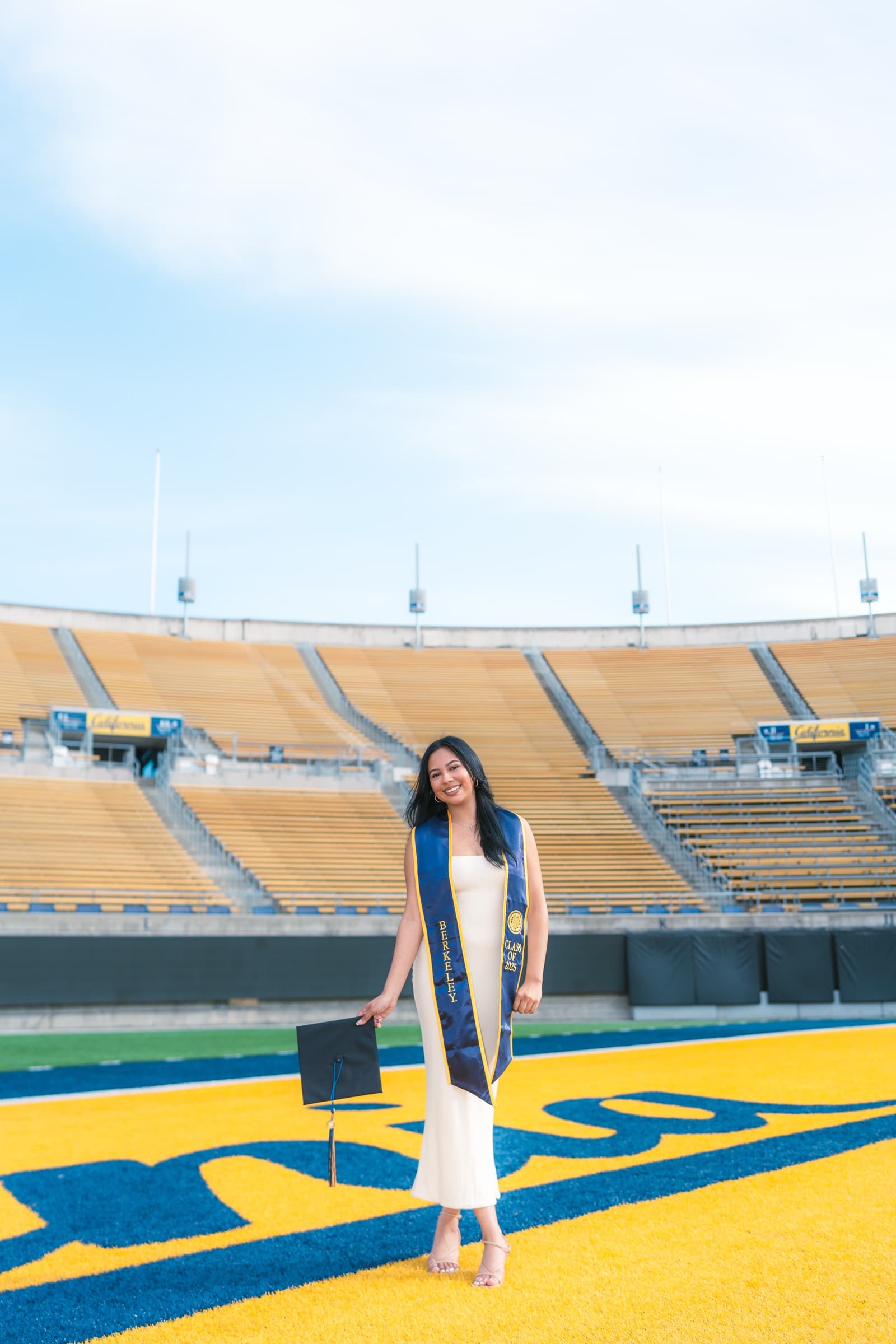 Graduate holding her cap on the field at California Memorial Stadium, empty gold bleachers behind